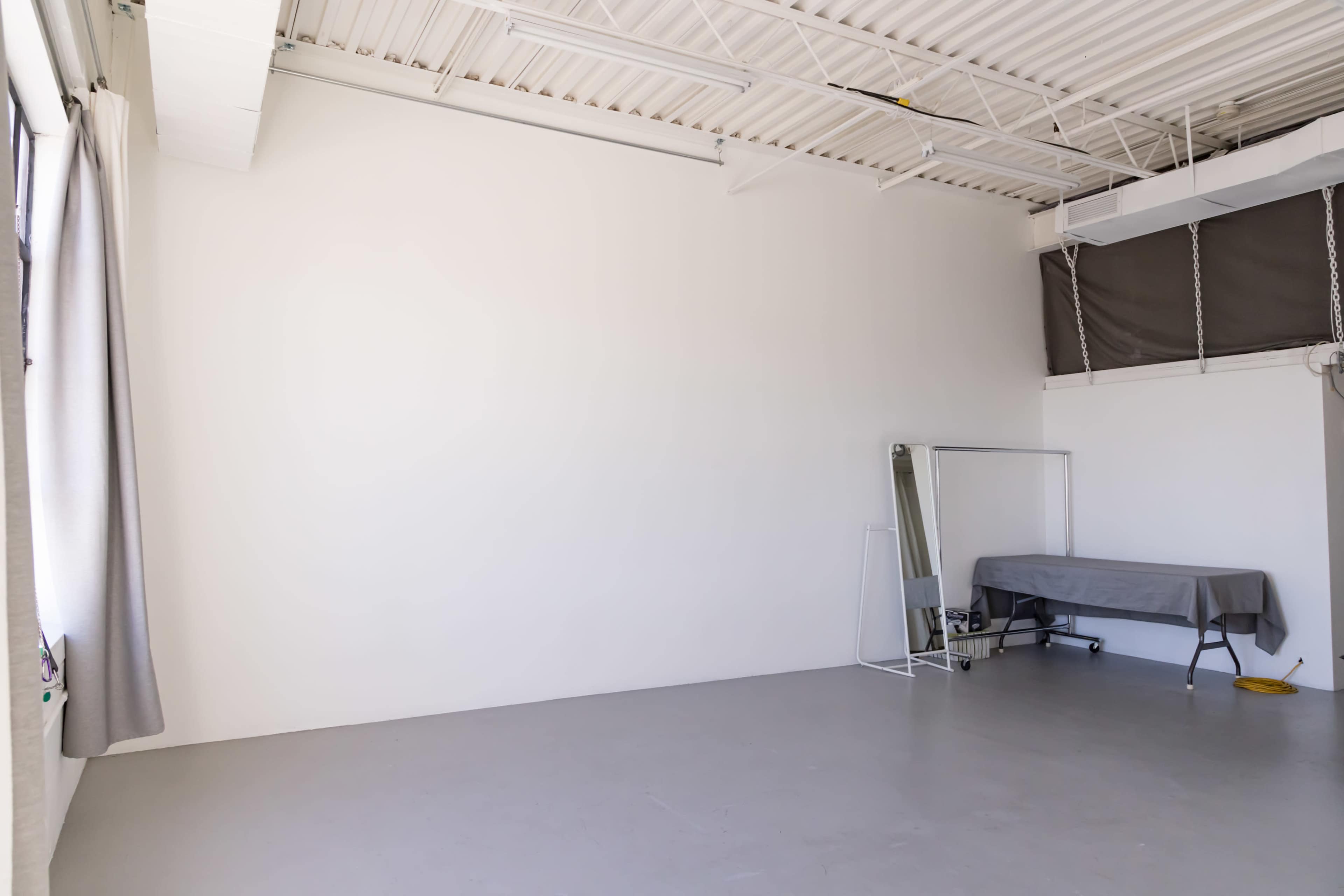 The image shows a minimalist room with a gray floor, empty white walls, a folding table covered by a tablecloth, a mirror, and natural light coming through a window.