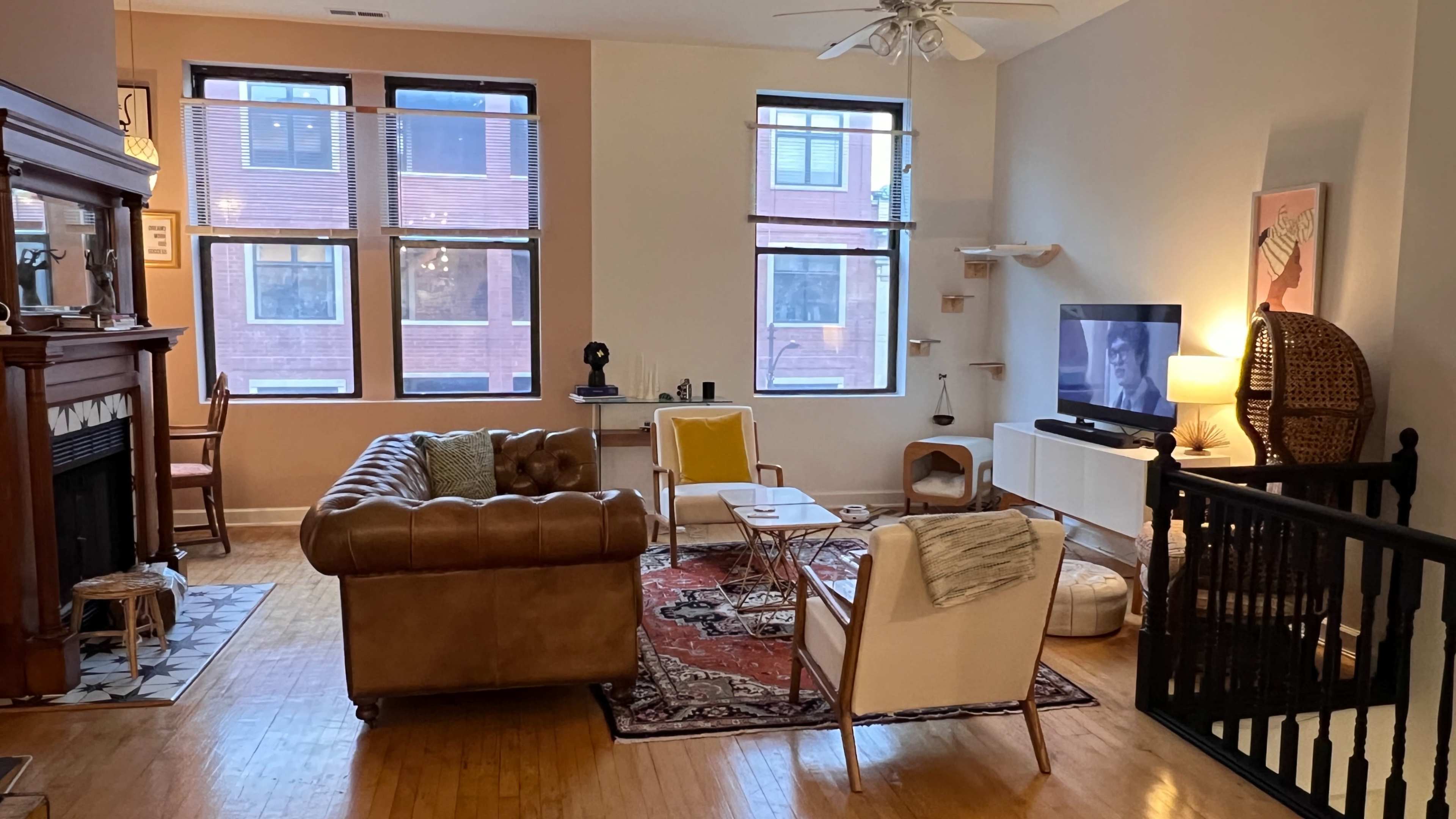 A spacious living room with large windows, a brown leather couch, a white media console with a television, and a mix of seating arrangements.