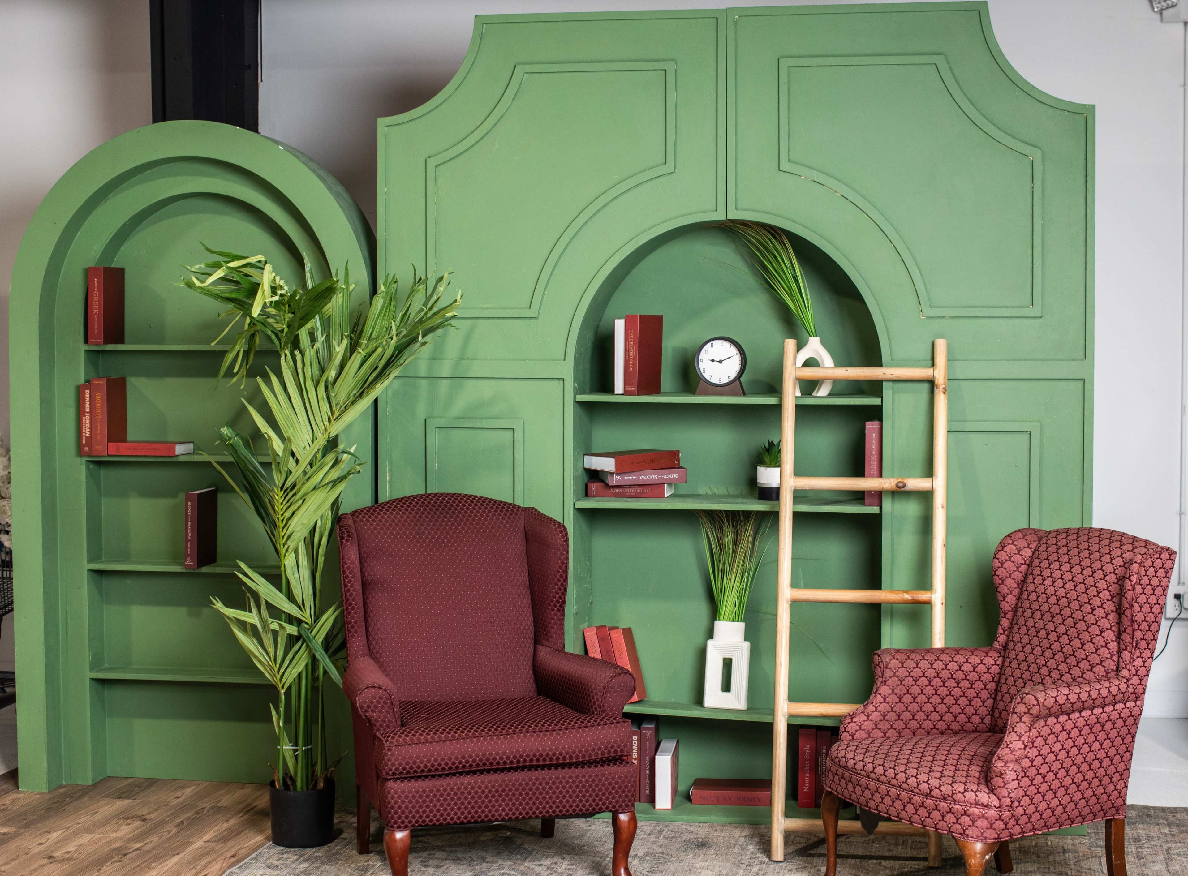 The image features a stylish indoor setup with two patterned armchairs flanking a green bookshelf backdrop adorned with plants, books, and a clock.