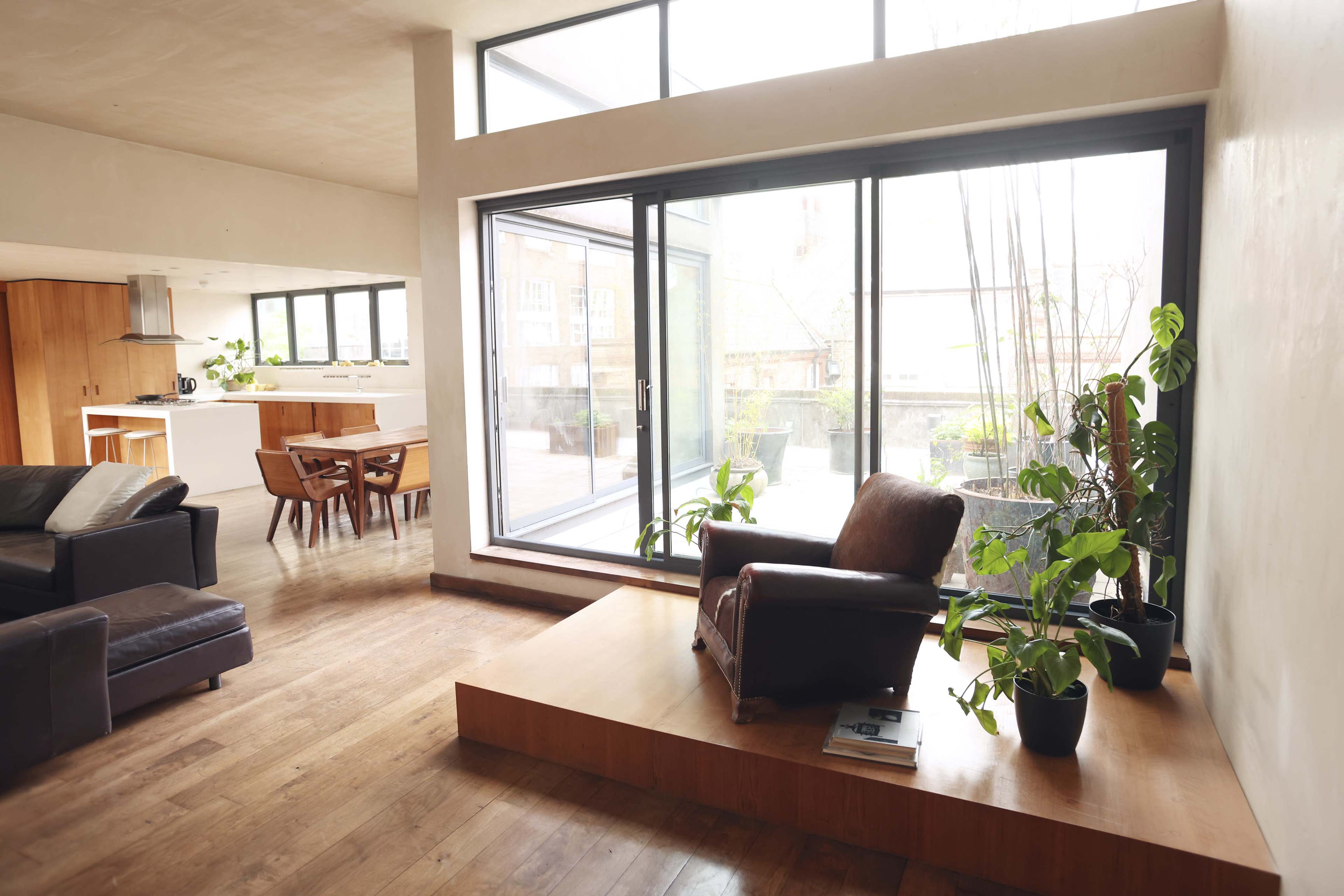 A spacious living area features a brown leather armchair on a raised wooden platform, with large windows providing natural light and a view of indoor plants.