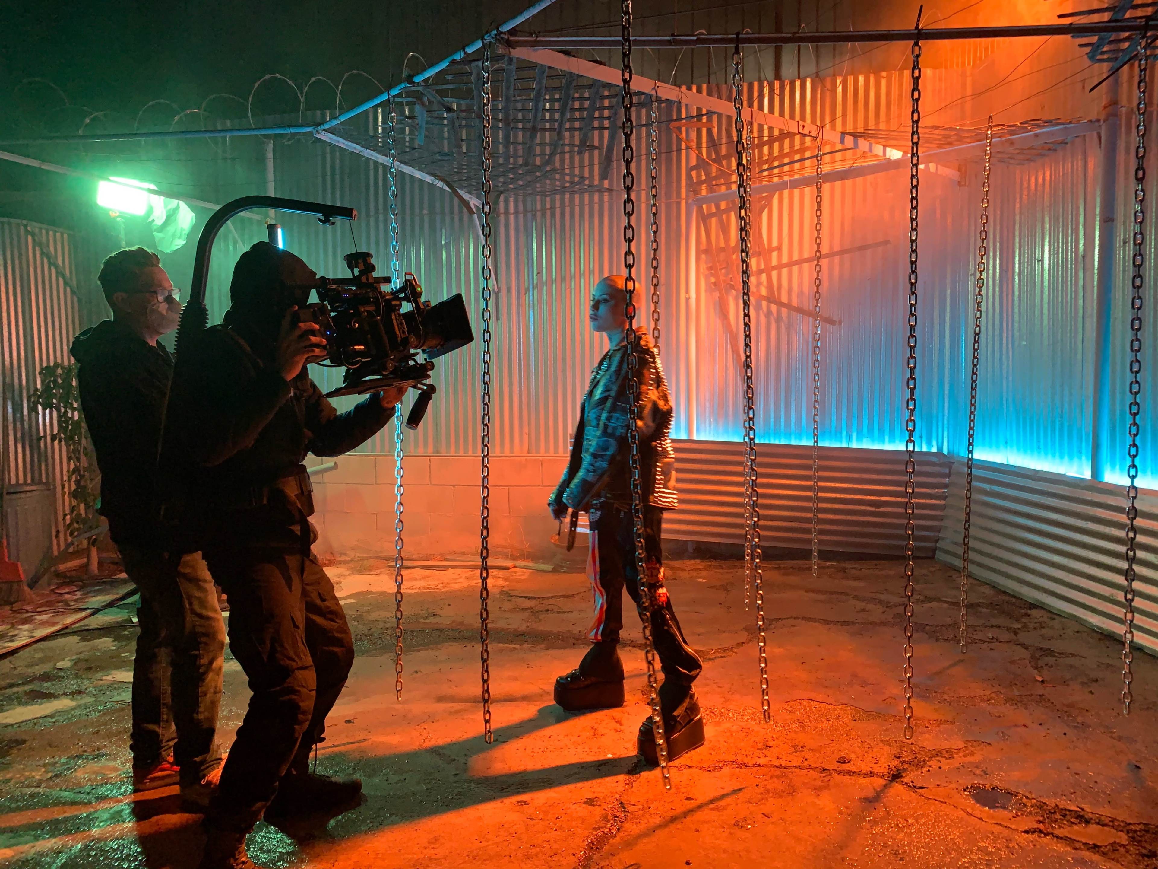 A film crew operates a camera on a set featuring a person walking among hanging chains and illuminated by colored lights.