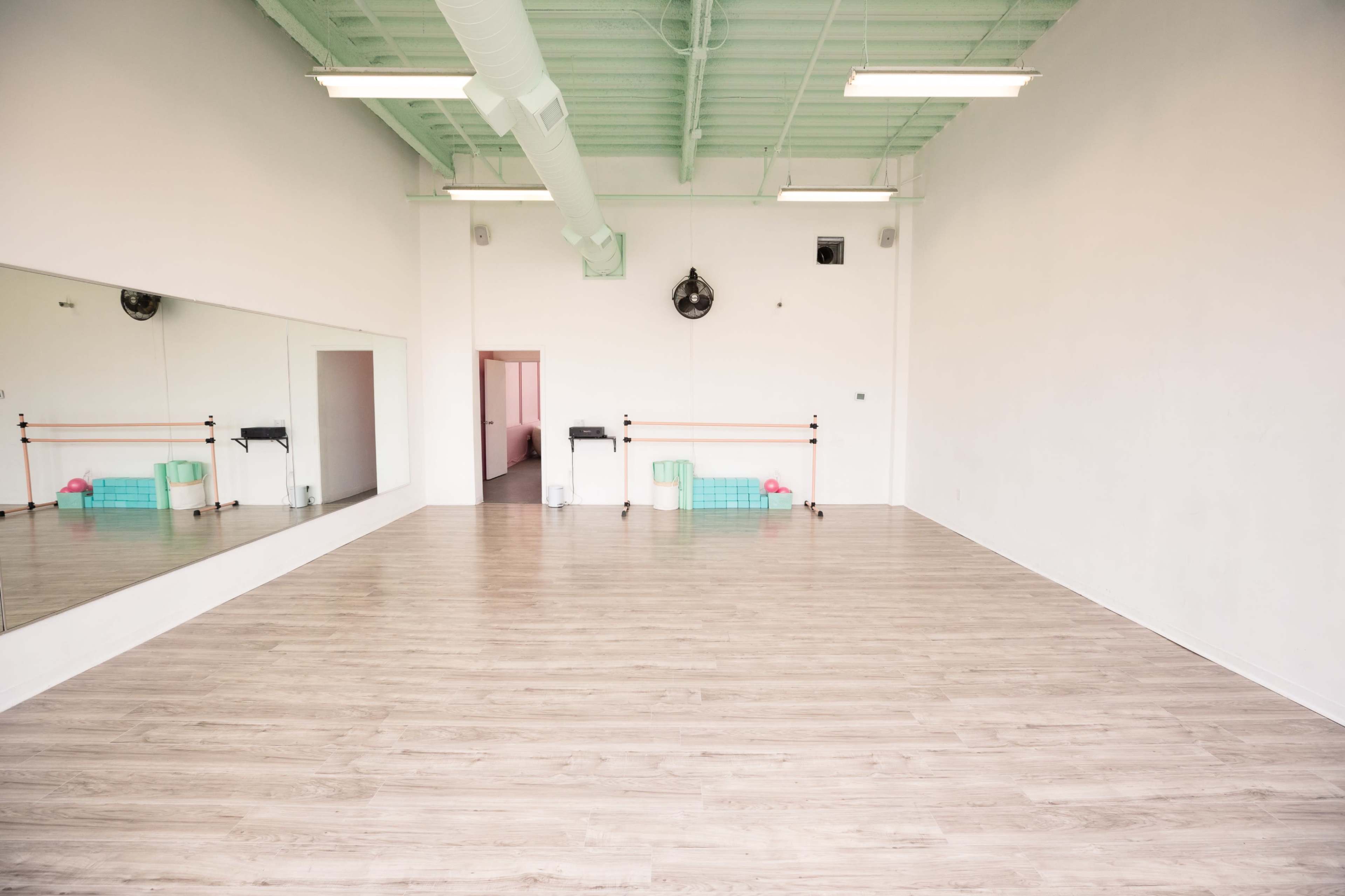 A spacious, empty room with wooden floors, a large mirror on one wall, and minimal furnishings along the back.