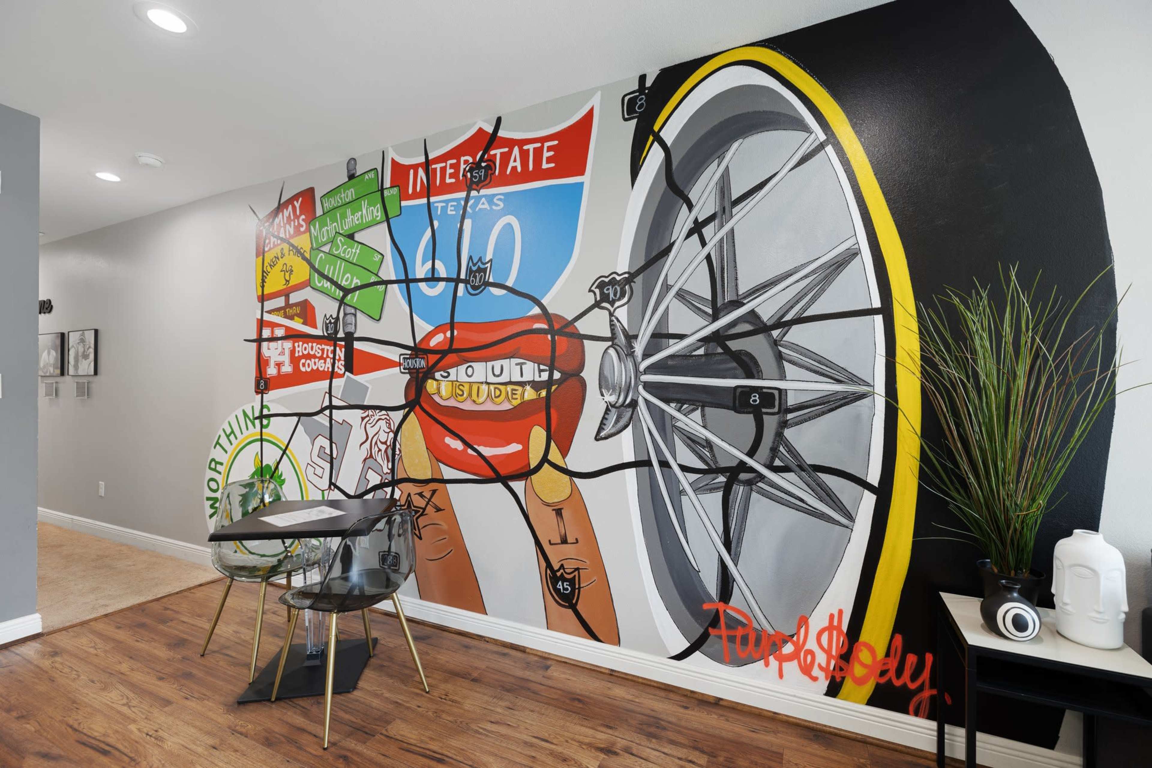 A dining area featuring a large mural of a bicycle wheel and various graphic elements on the wall, alongside a glass table and chairs.