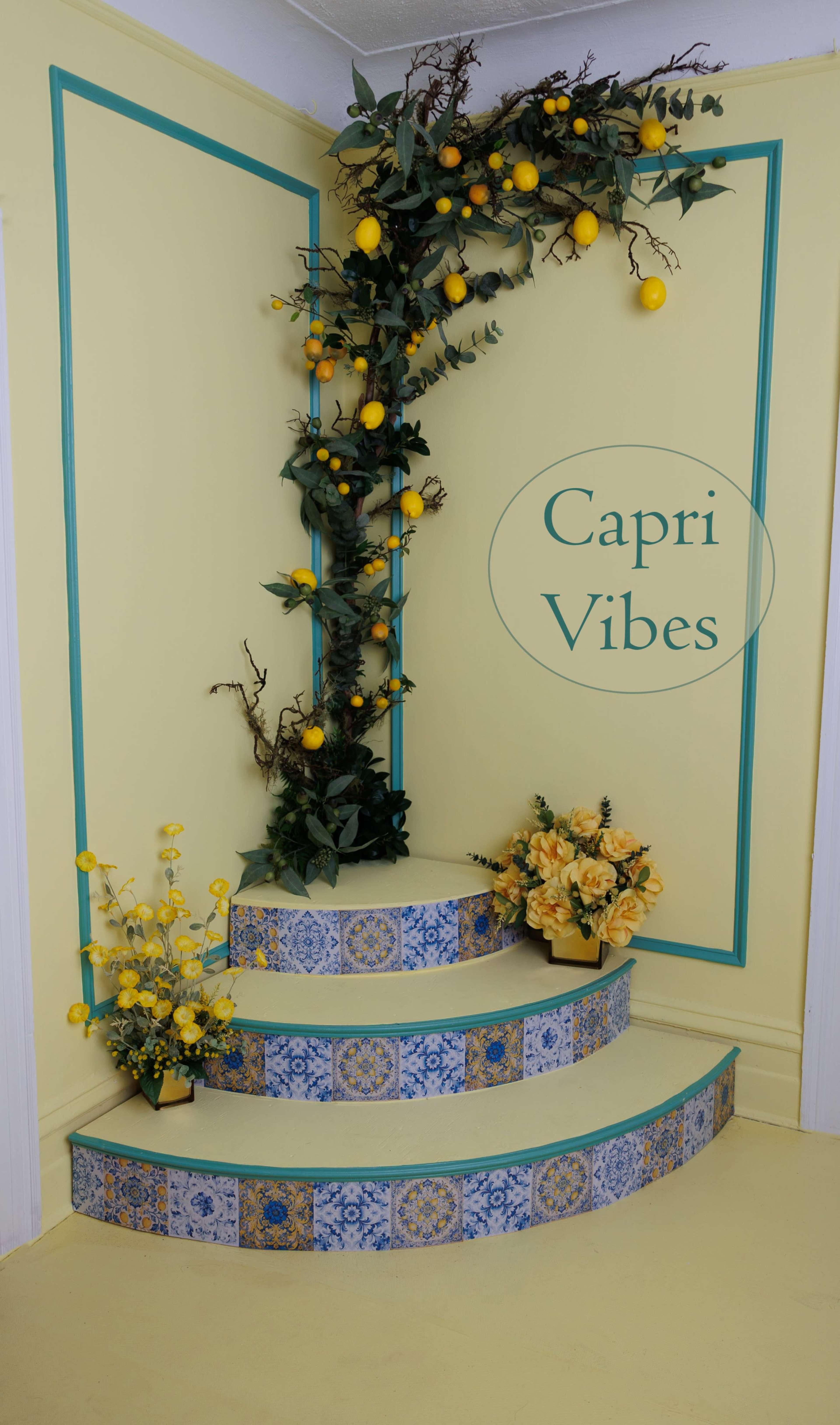 The image shows a decorative corner with a spiral staircase featuring blue and yellow tiles, adorned with yellow flowers and foliage, set against a light yellow wall.