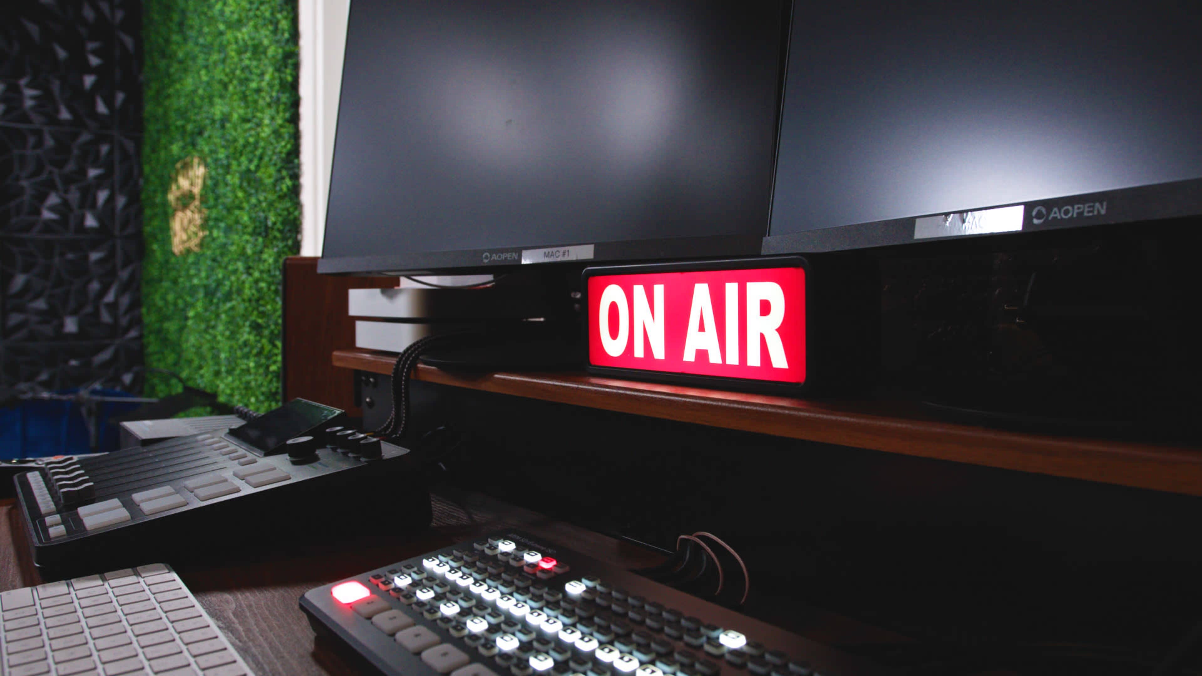 A red "ON AIR" sign is illuminated near two computer monitors on a broadcast studio desk, alongside control equipment.