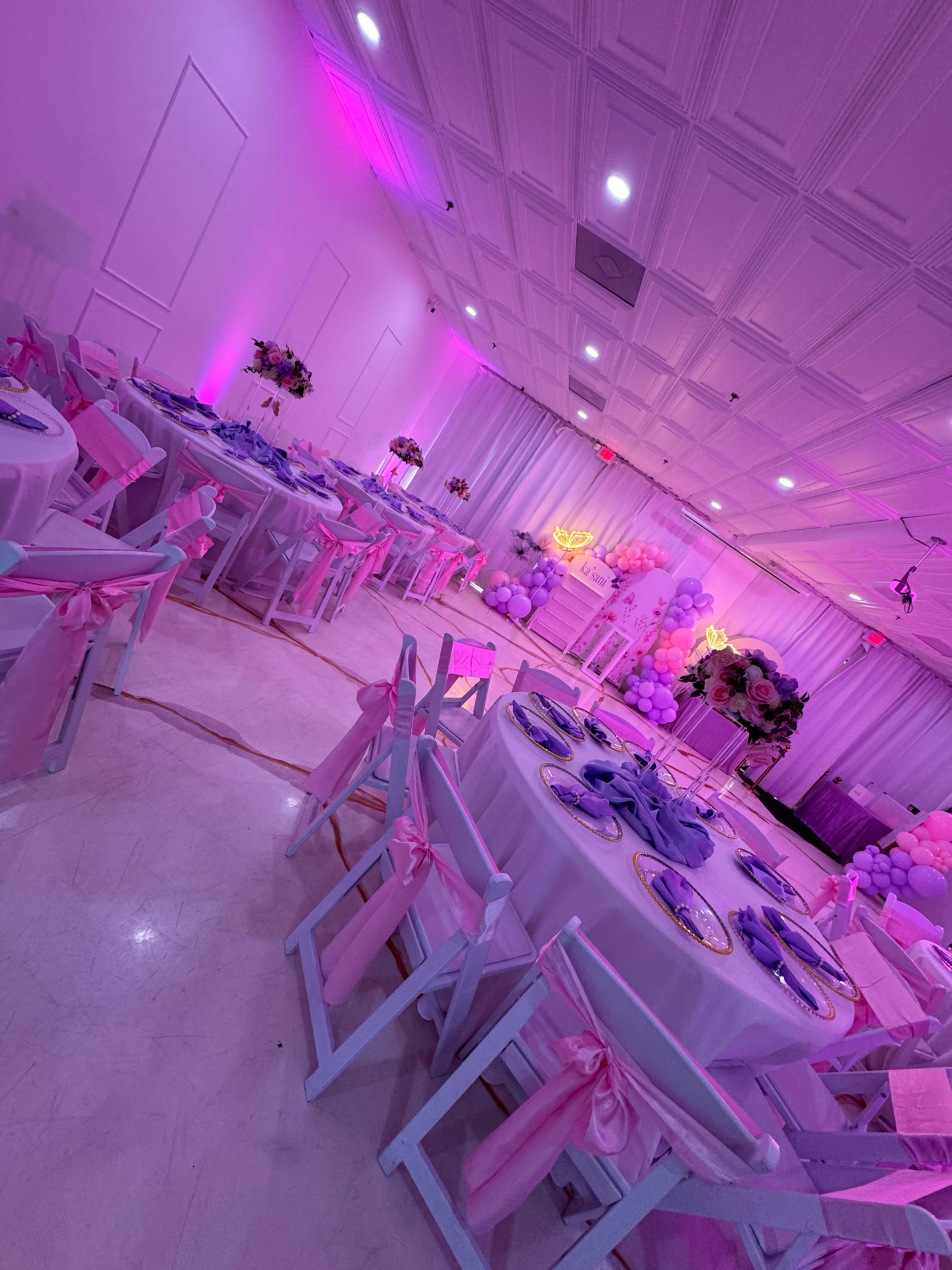 The image shows a decorated event space with tables set with blue plates, pink tablecloths, and white chairs adorned with pink bows, illuminated by pink lighting.