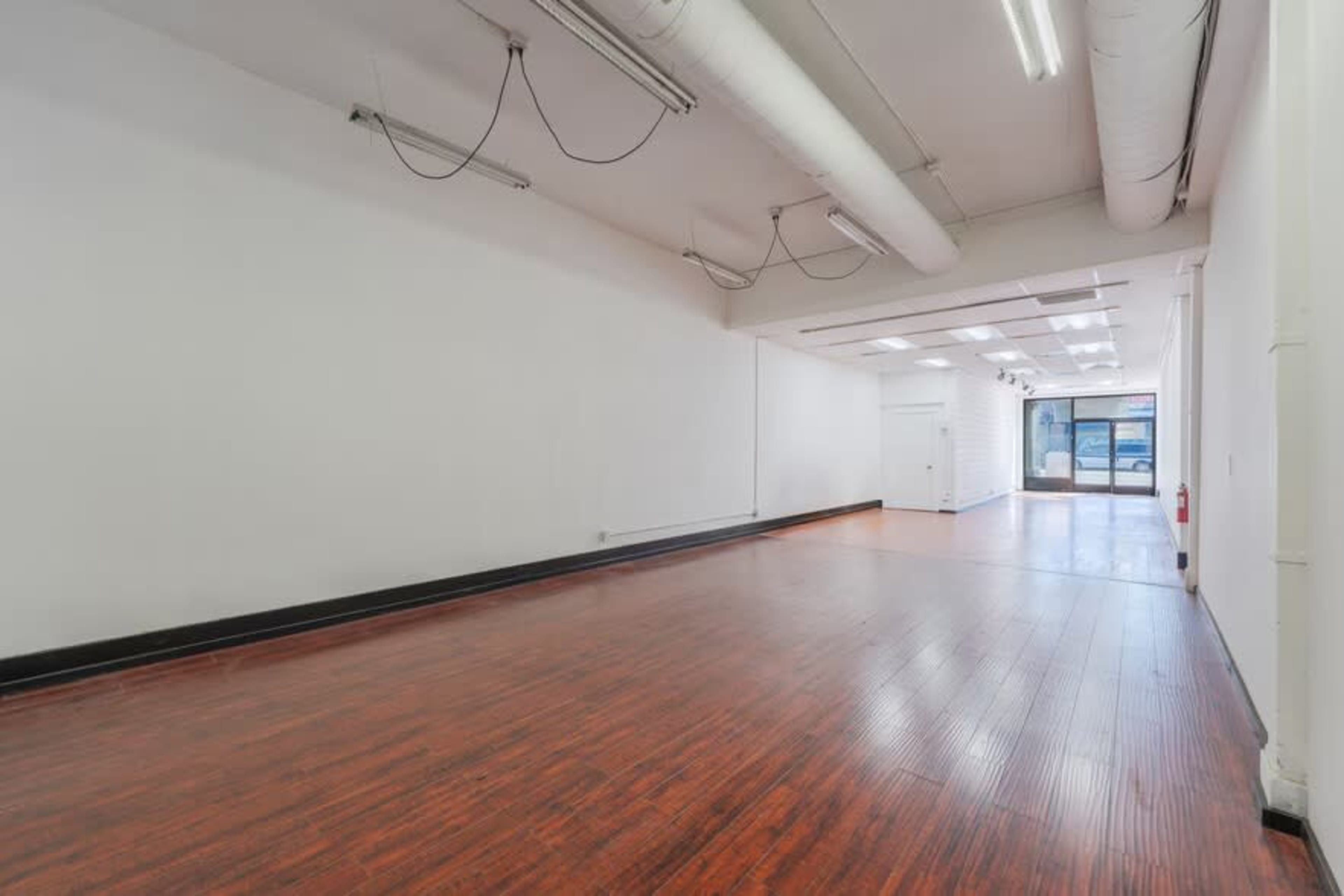 The image shows an empty commercial space with wooden flooring, white walls, and large windows at the front.