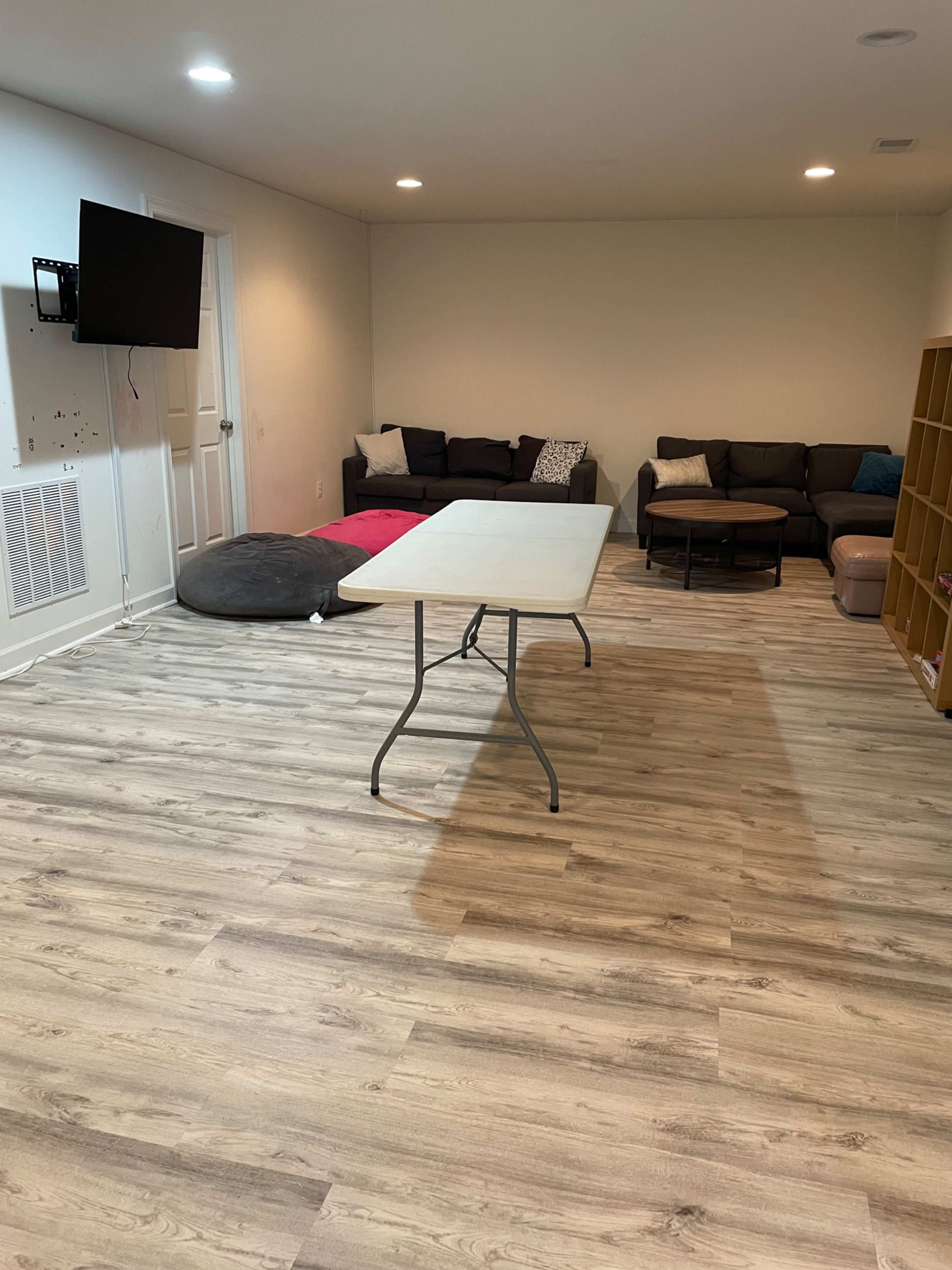 The image shows a spacious room with a large table in the center, a couch along one wall, a bean bag chair, and a shelving unit against another wall.