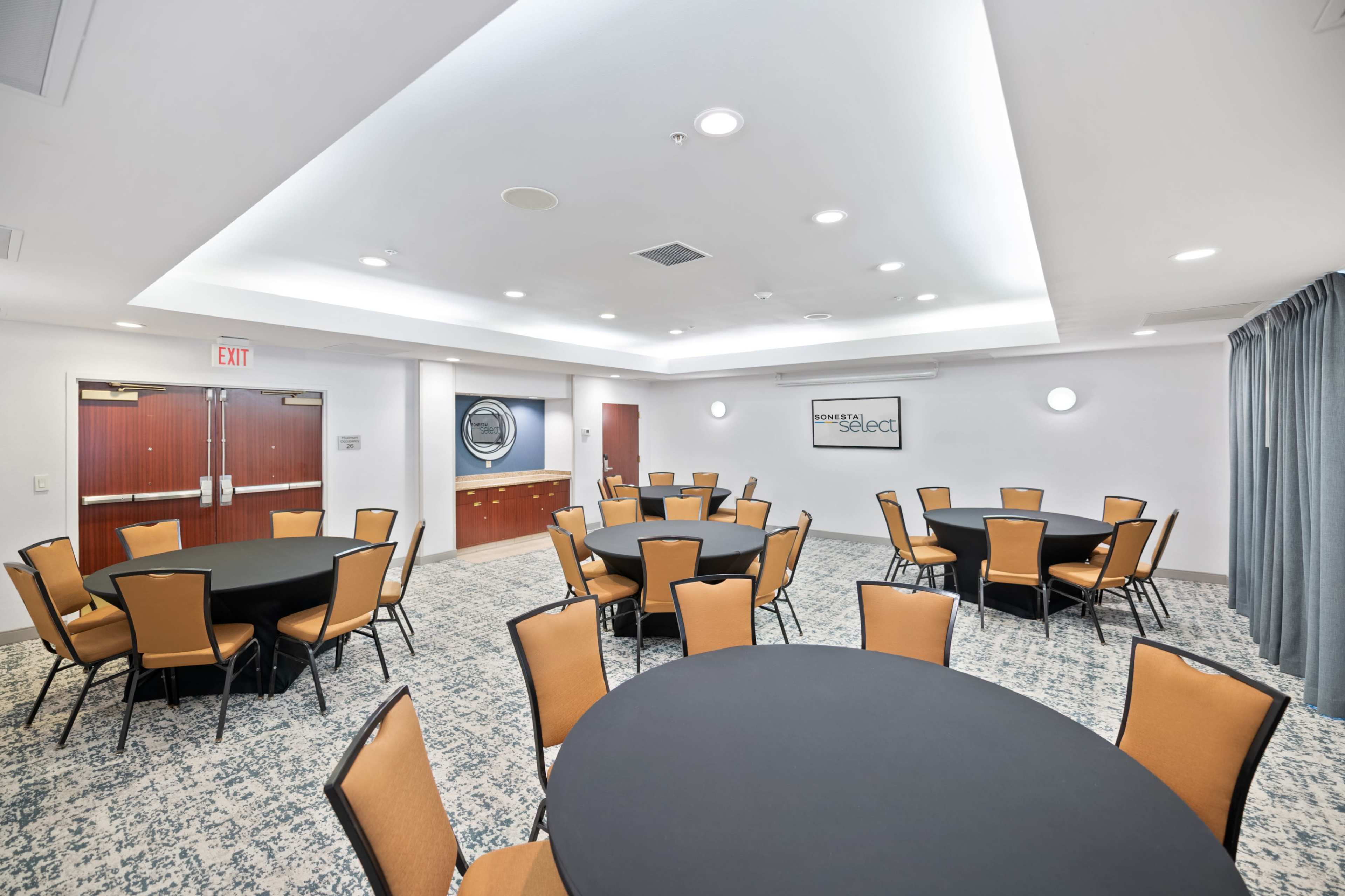 The image shows a modern meeting room with several round tables surrounded by chairs, featuring a gray carpet and well-lit ceiling.
