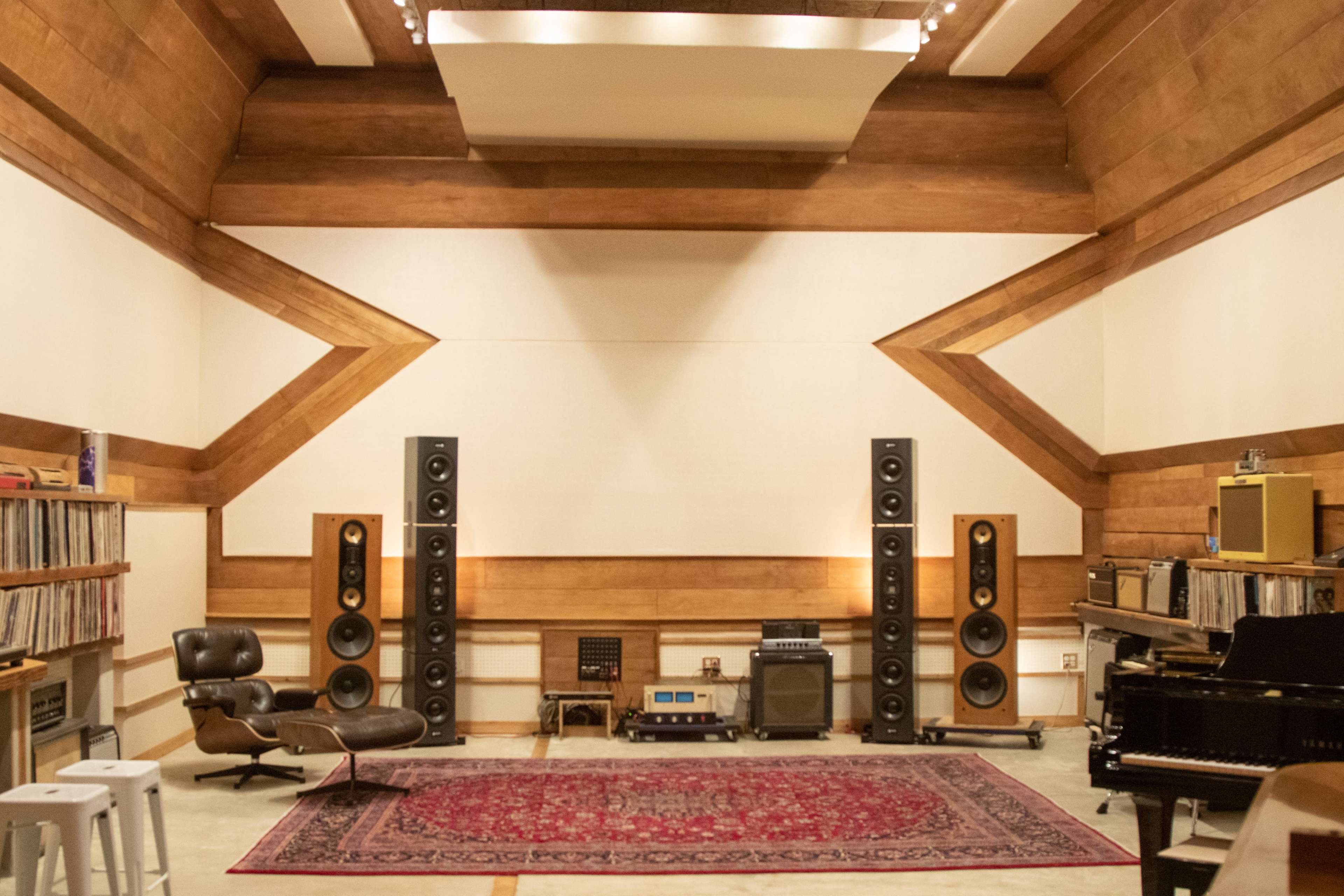 A music studio with tall speakers on either side, a piano, wooden walls, and shelves filled with vinyl records.