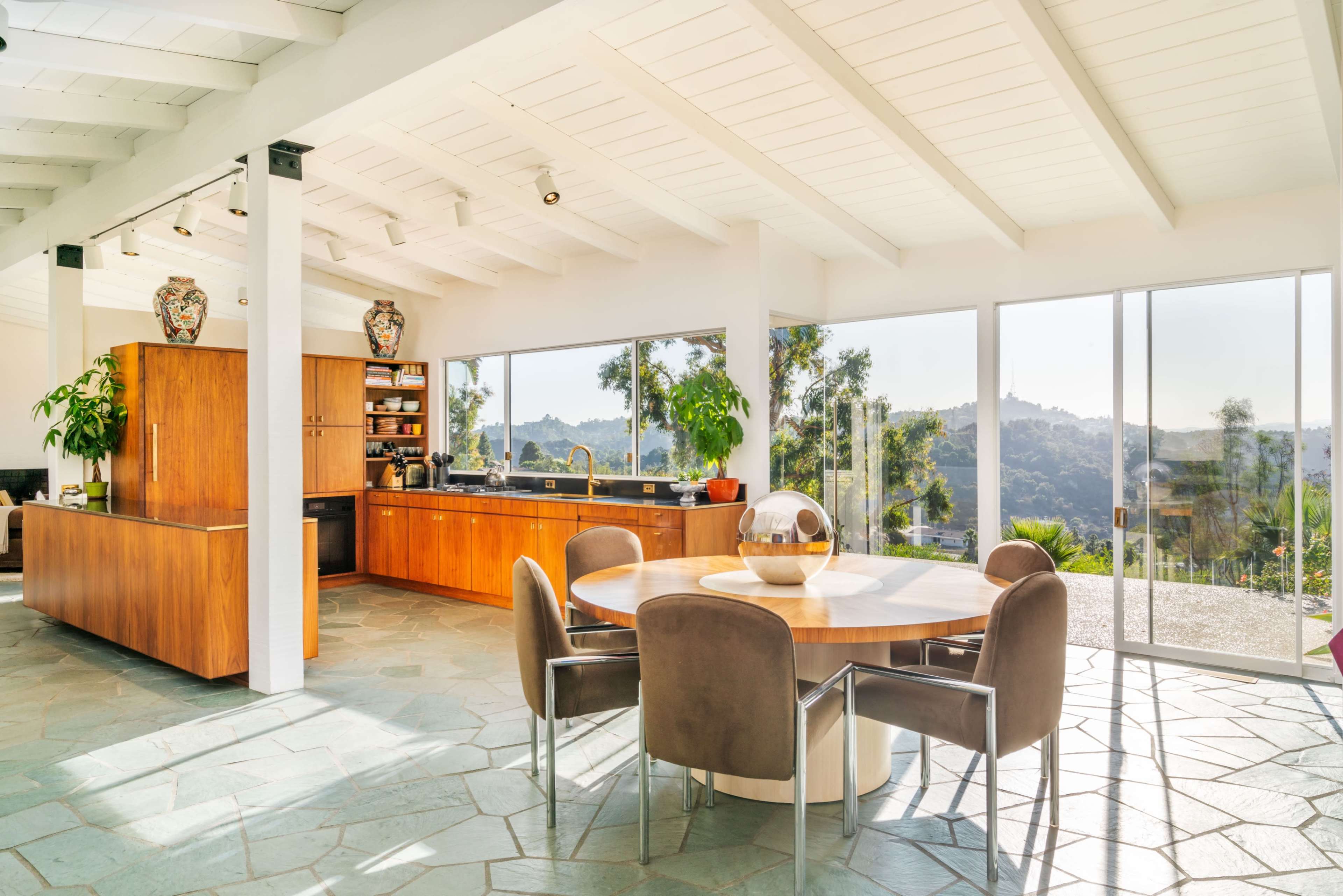 A modern kitchen and dining area with wooden cabinetry and a round dining table, featuring large windows that offer a view of the surrounding hills.
