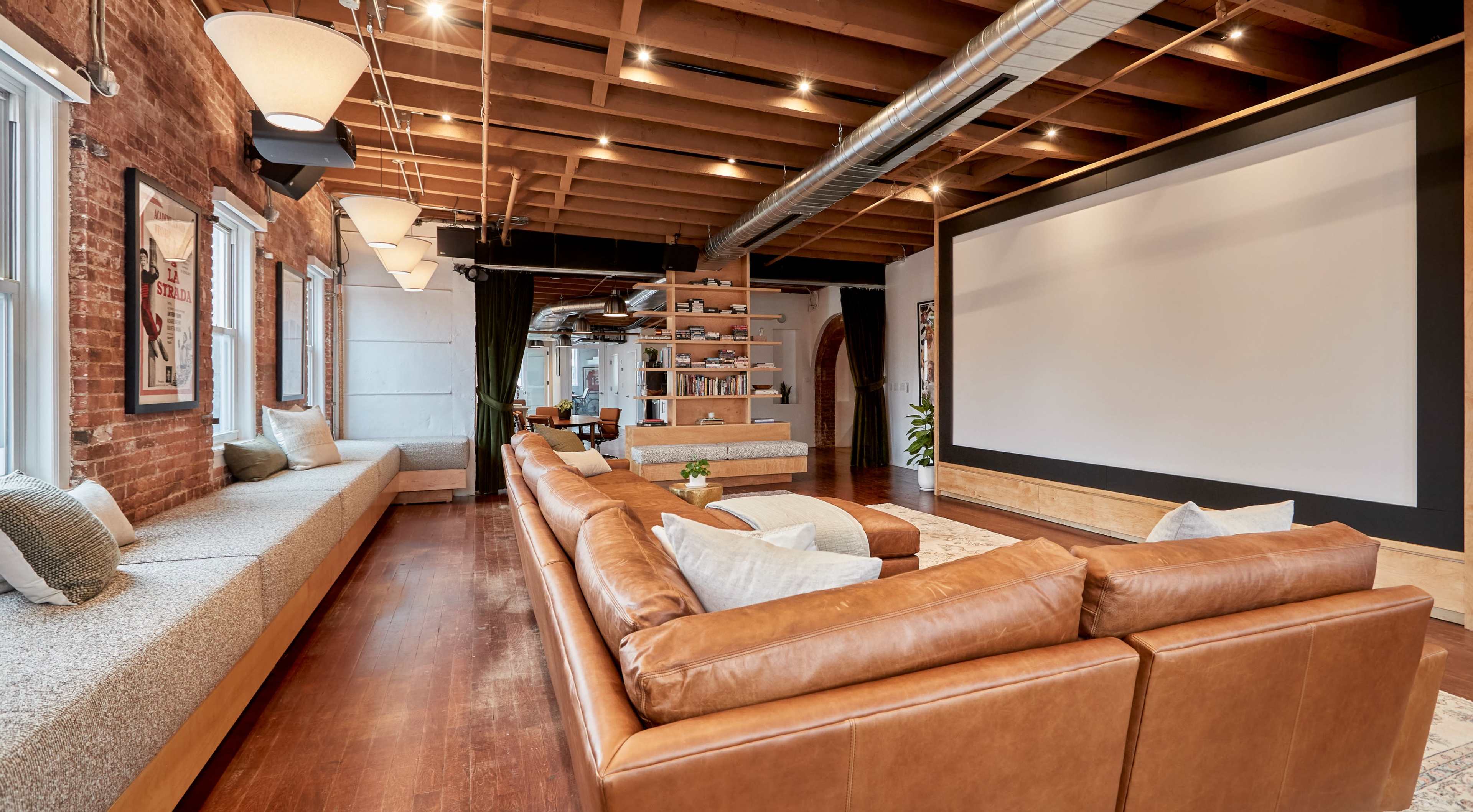 A spacious living room featuring a large brown leather sectional sofa, a wall-mounted screen, and a cozy seating area beneath windows with wooden beams overhead.