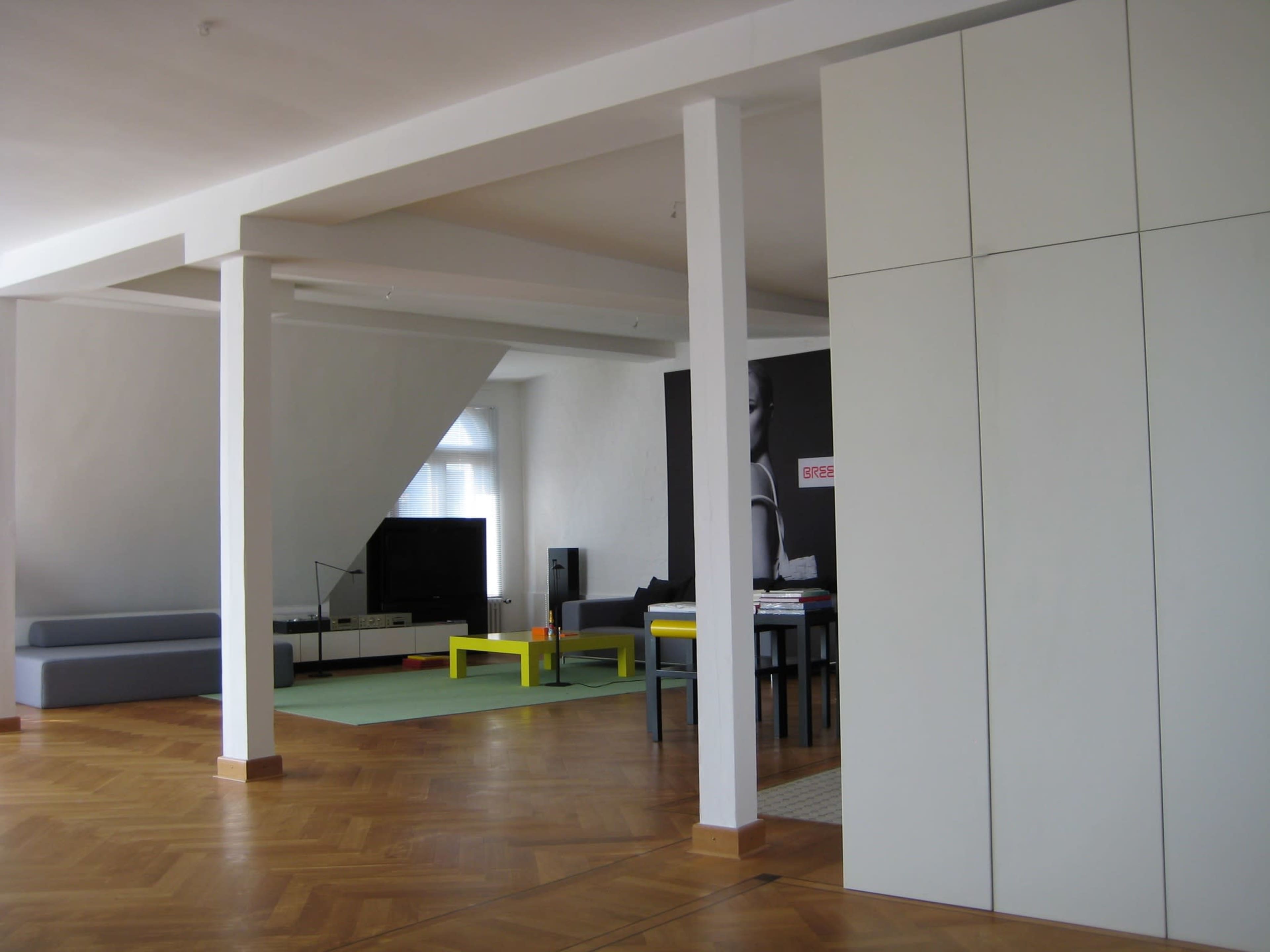 The image shows a spacious, modern living area with a sectional sofa, a television, a dining table with colorful chairs, and large windows.