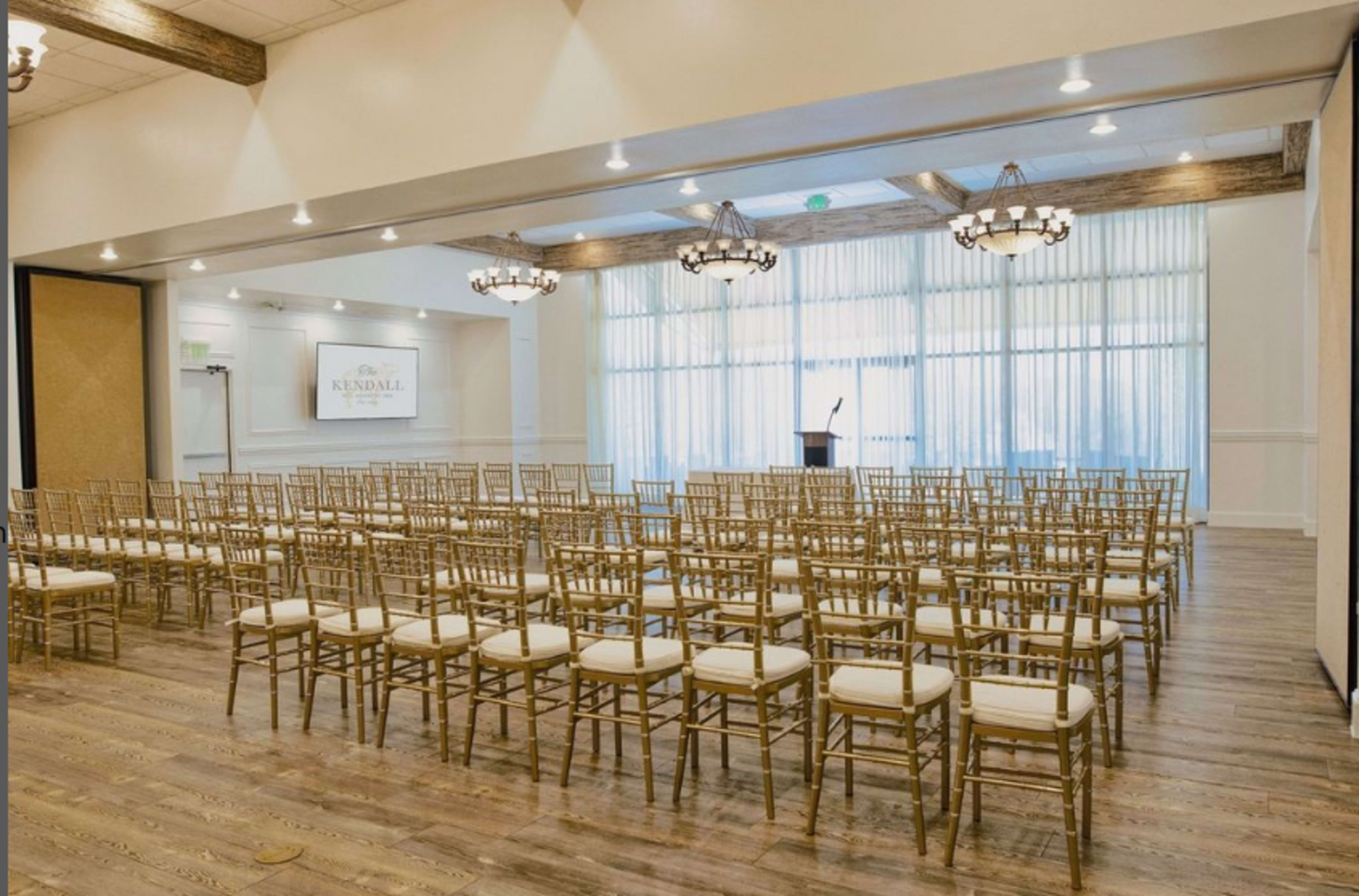 The image shows a conference room set up with rows of gold chiavari chairs facing a podium under decorative chandeliers.