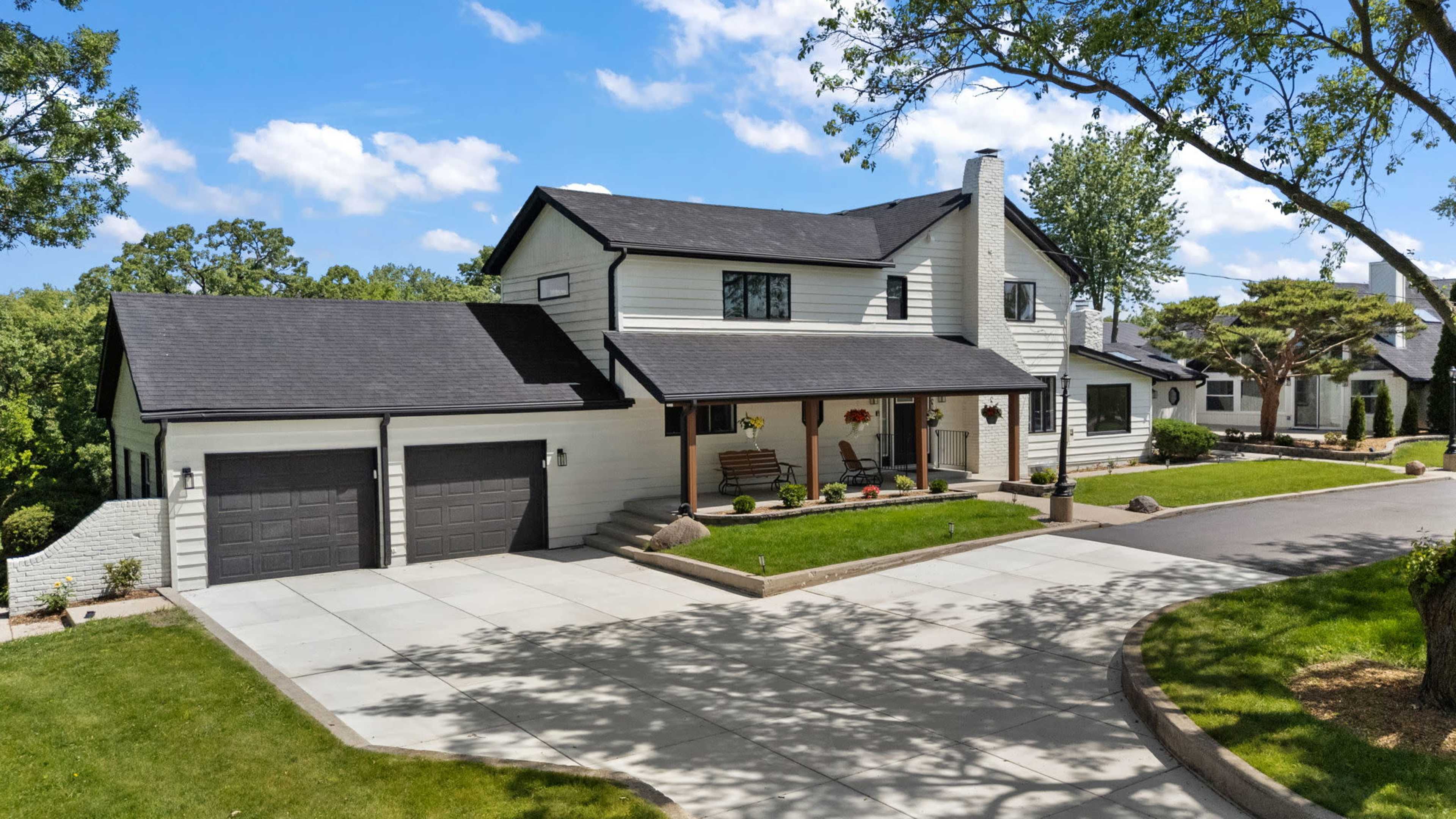 The image shows a modern two-story house with a driveway, front lawn, and garage doors on a sunny day.
