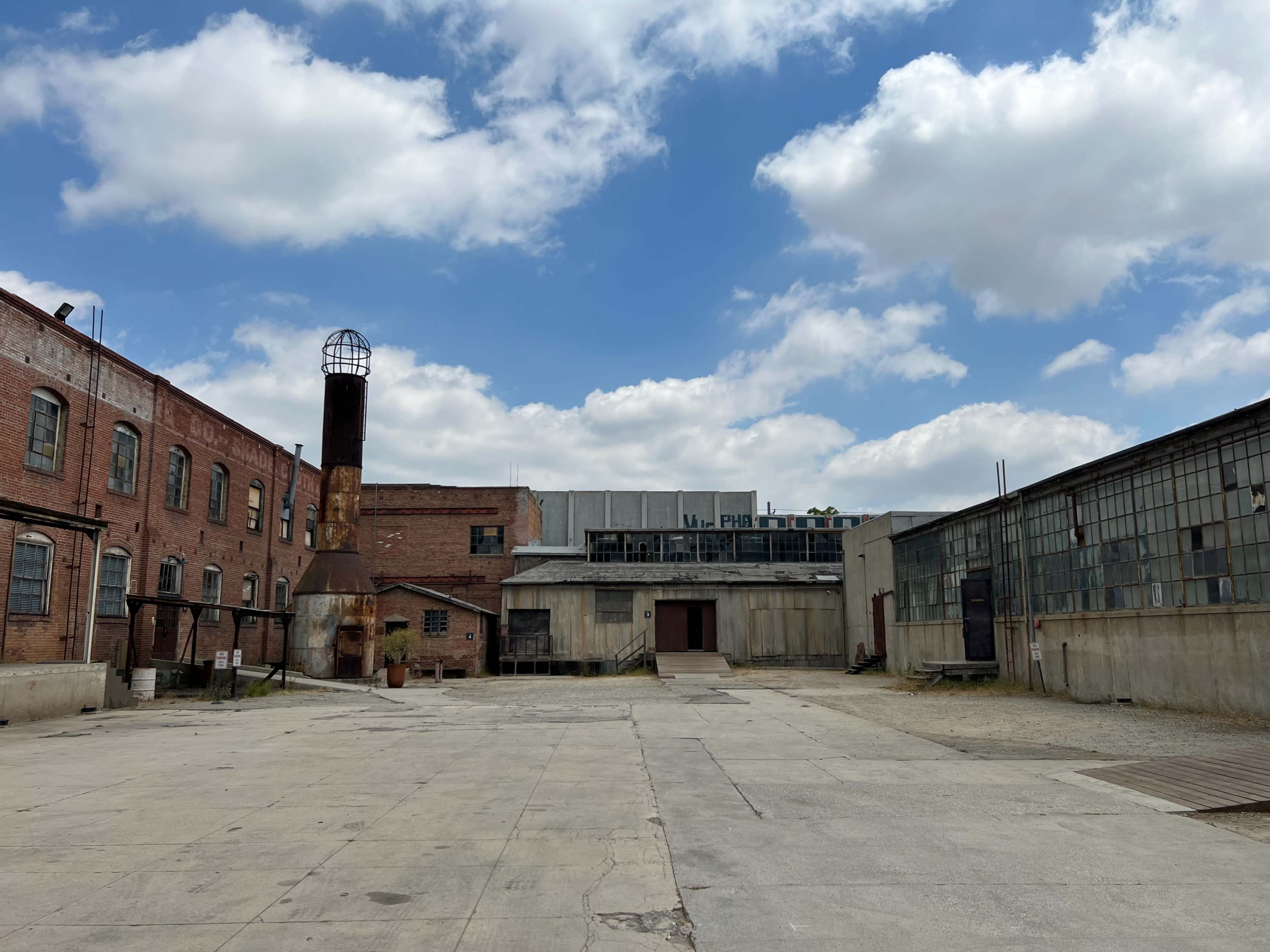 An industrial complex with brick buildings, a chimney, and a large open yard under a cloudy sky.