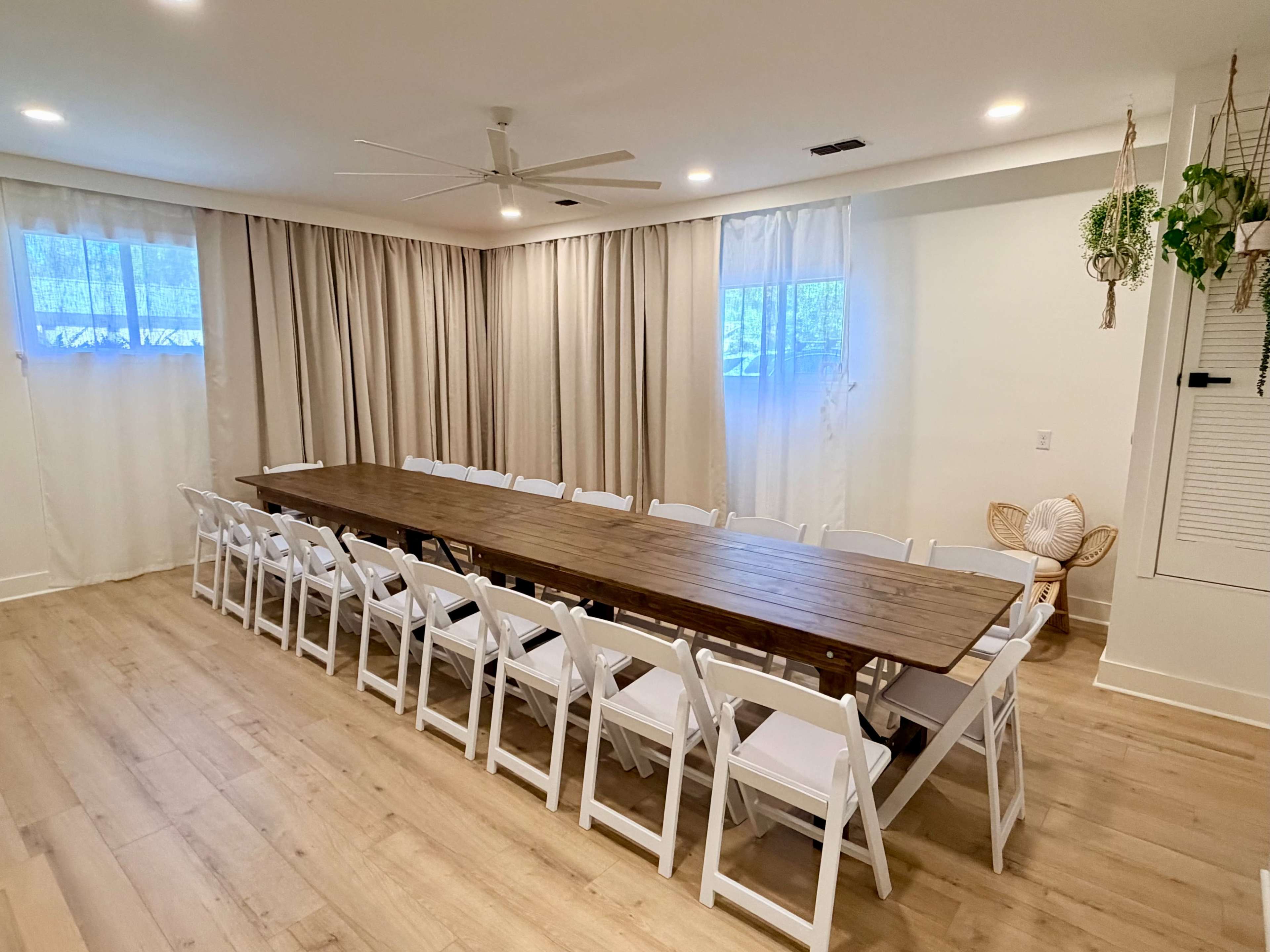 The image shows a spacious meeting room with a long wooden table surrounded by white folding chairs, large windows, and light-colored curtains.