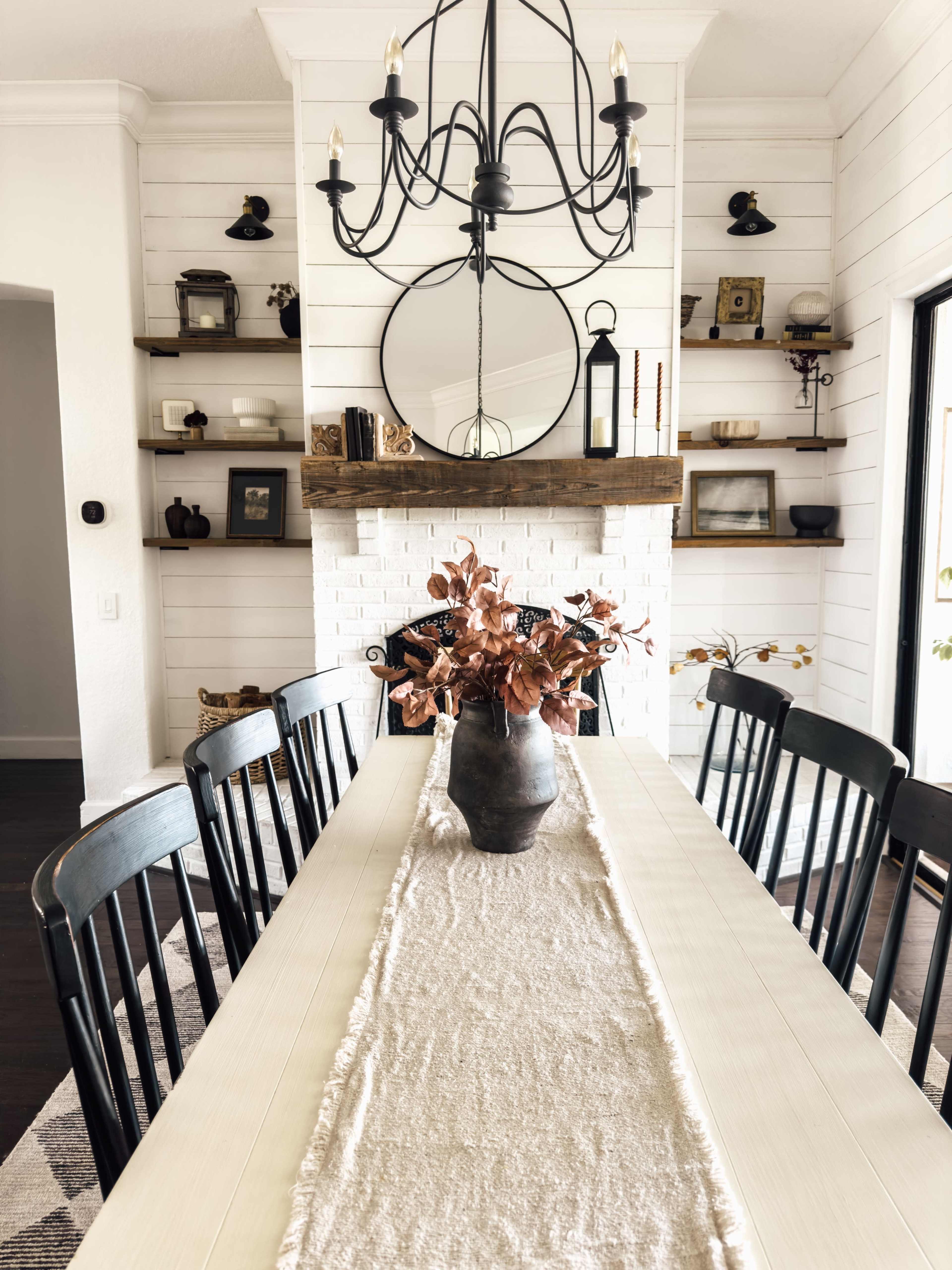 A modern dining room features a long table with black chairs, a centerpiece of dried plants, and a light fixture above, with shelves displaying decorative items on the walls.