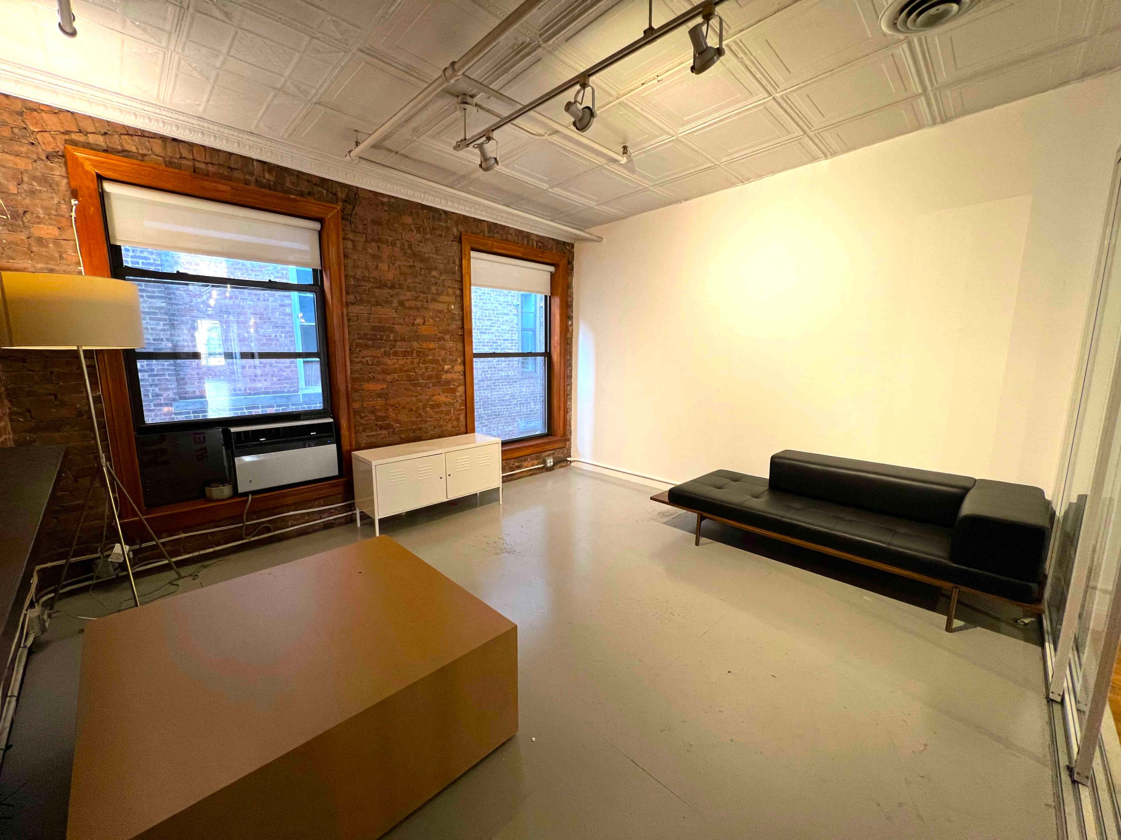 The image shows a minimalistic room with exposed brick walls, a black sofa, a light-colored coffee table, and a window air conditioning unit.