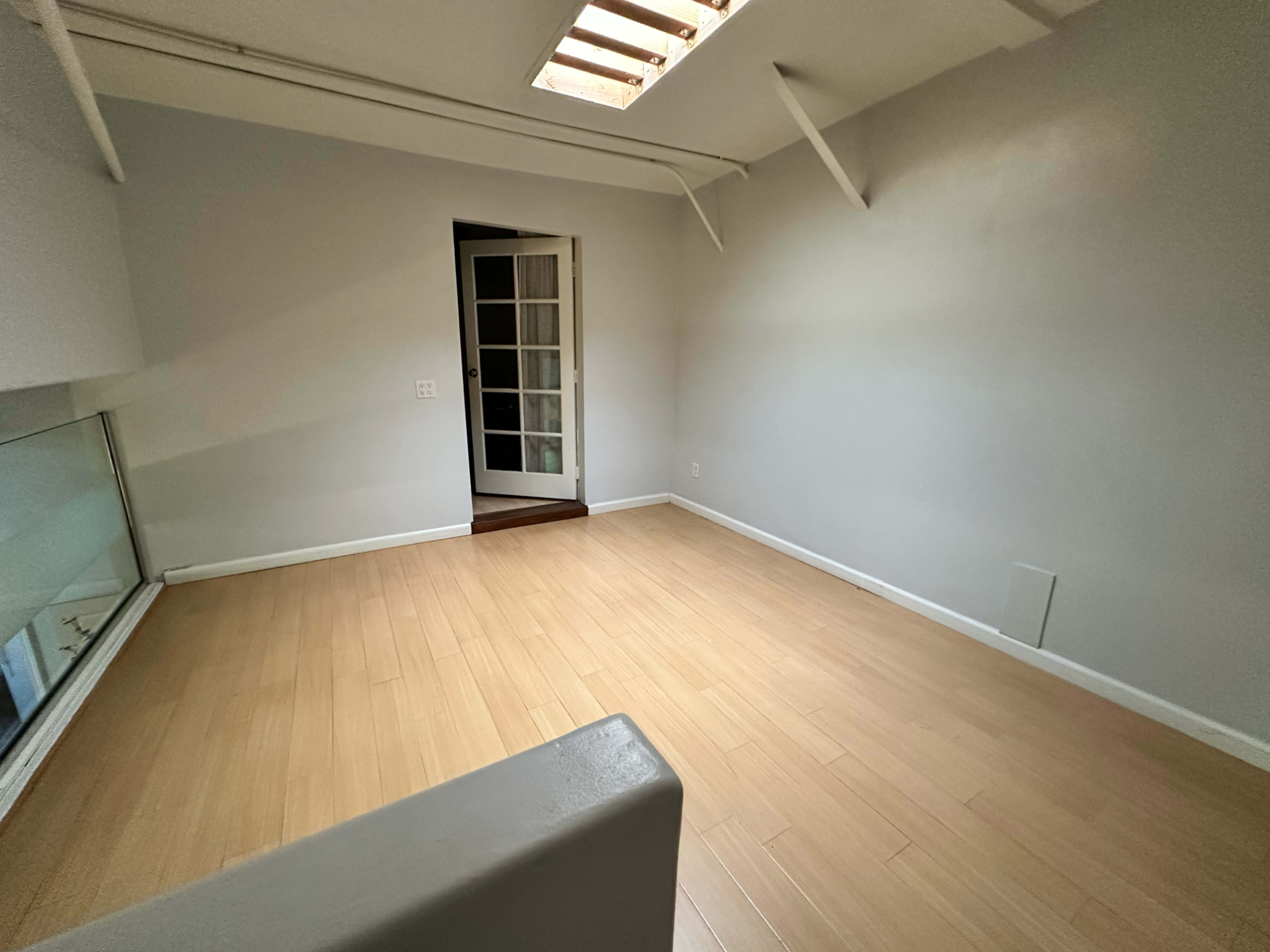 The image shows a vacant room with light-colored wooden flooring, white walls, and a doorway leading to another room.