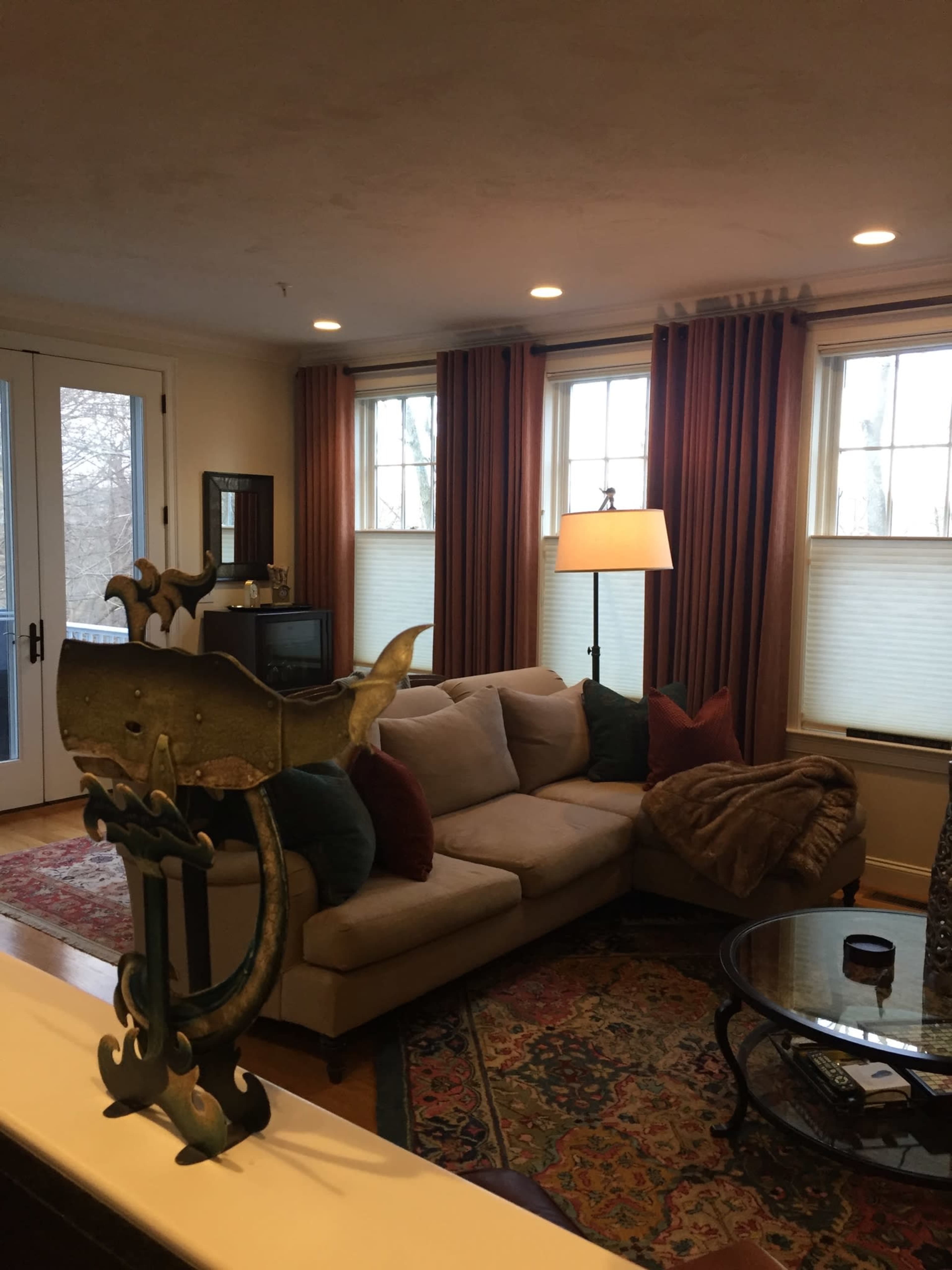 A well-lit living room features a beige sectional sofa with colorful pillows, a glass coffee table, and large windows dressed with curtains.