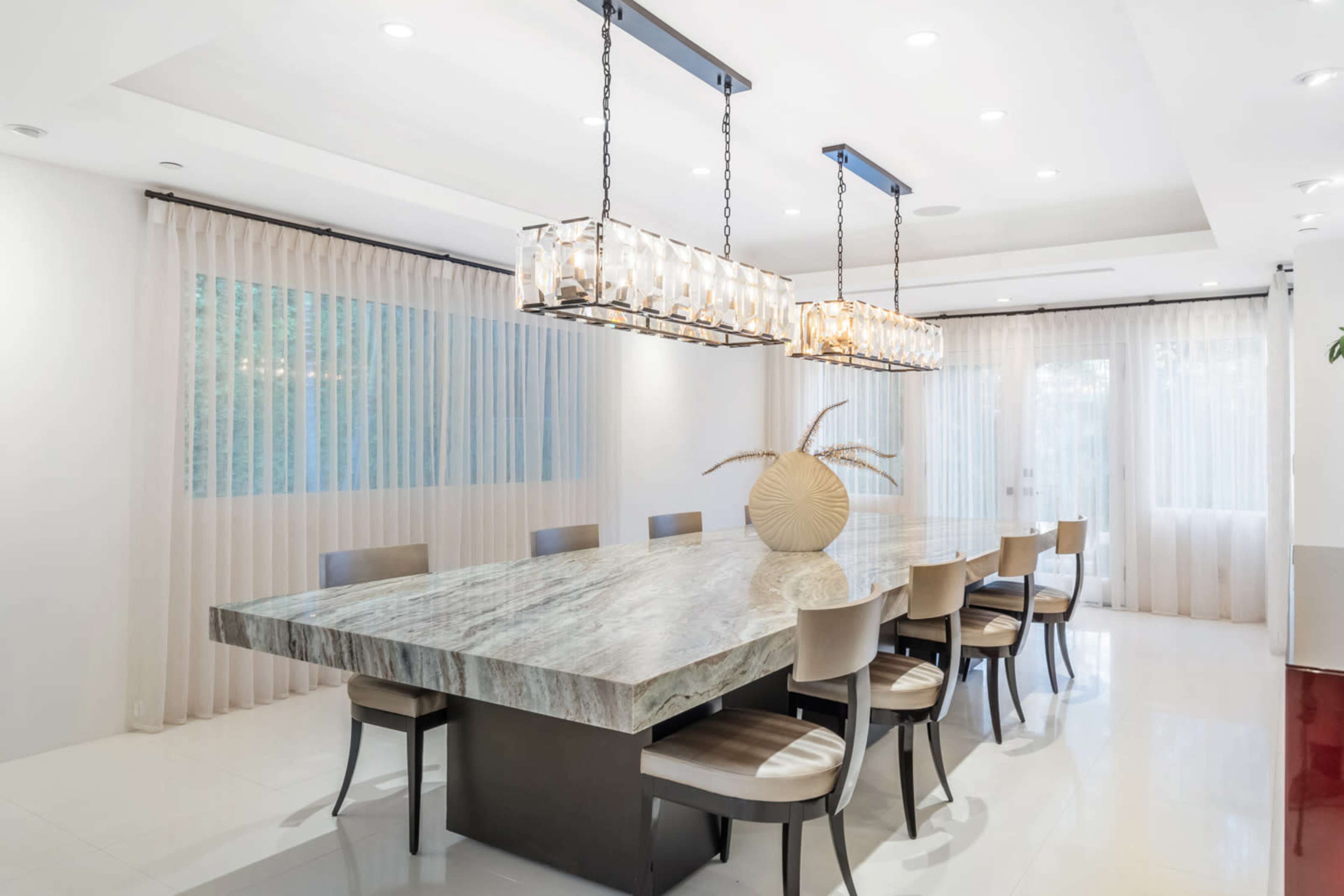 A modern dining room with a large marble table surrounded by eight chairs beneath two pendant light fixtures.