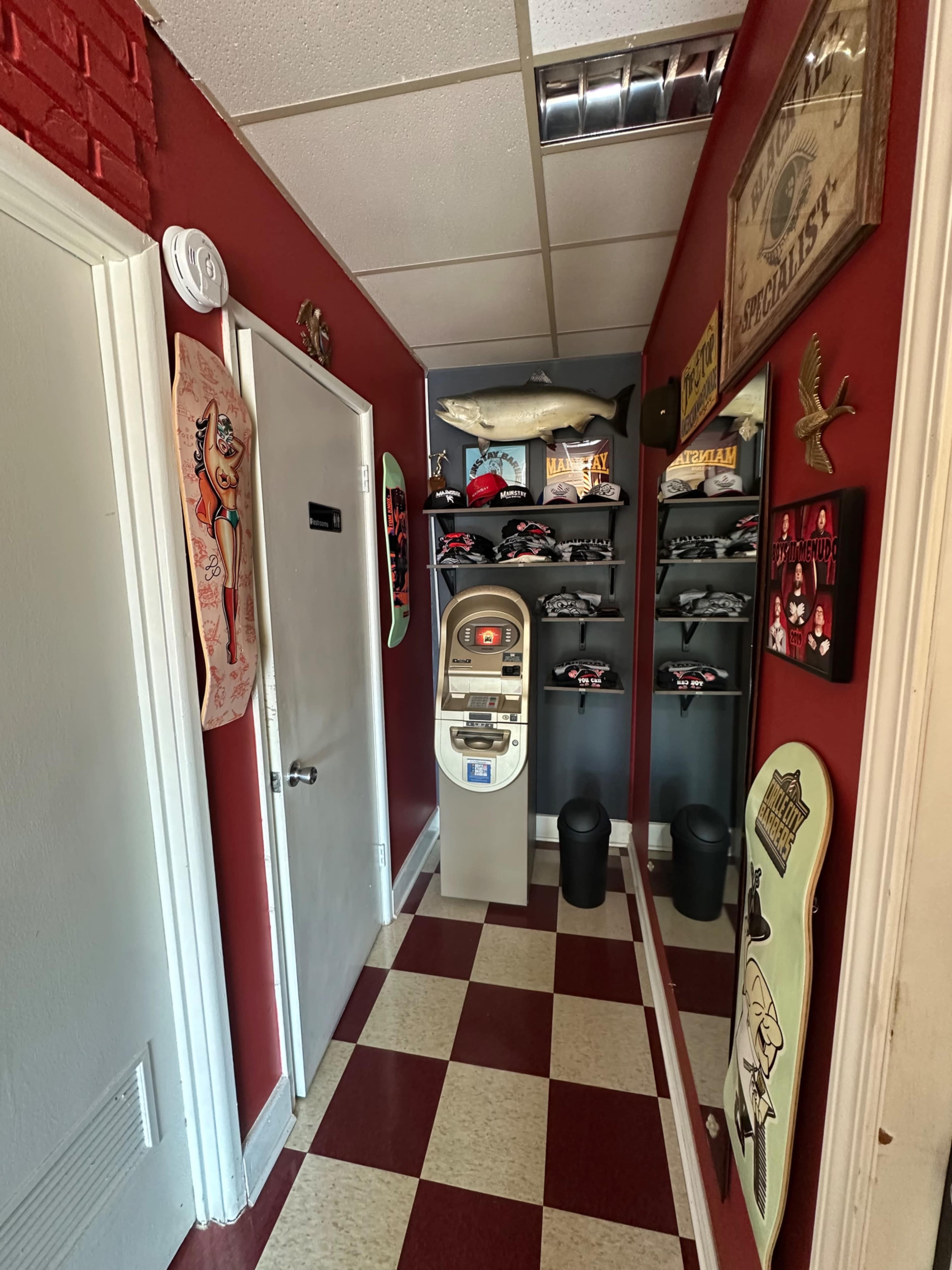 The image shows a narrow hallway with red walls, featuring various decorations, including a mounted fish, posters, helmets, and a vending machine, alongside checkered flooring.