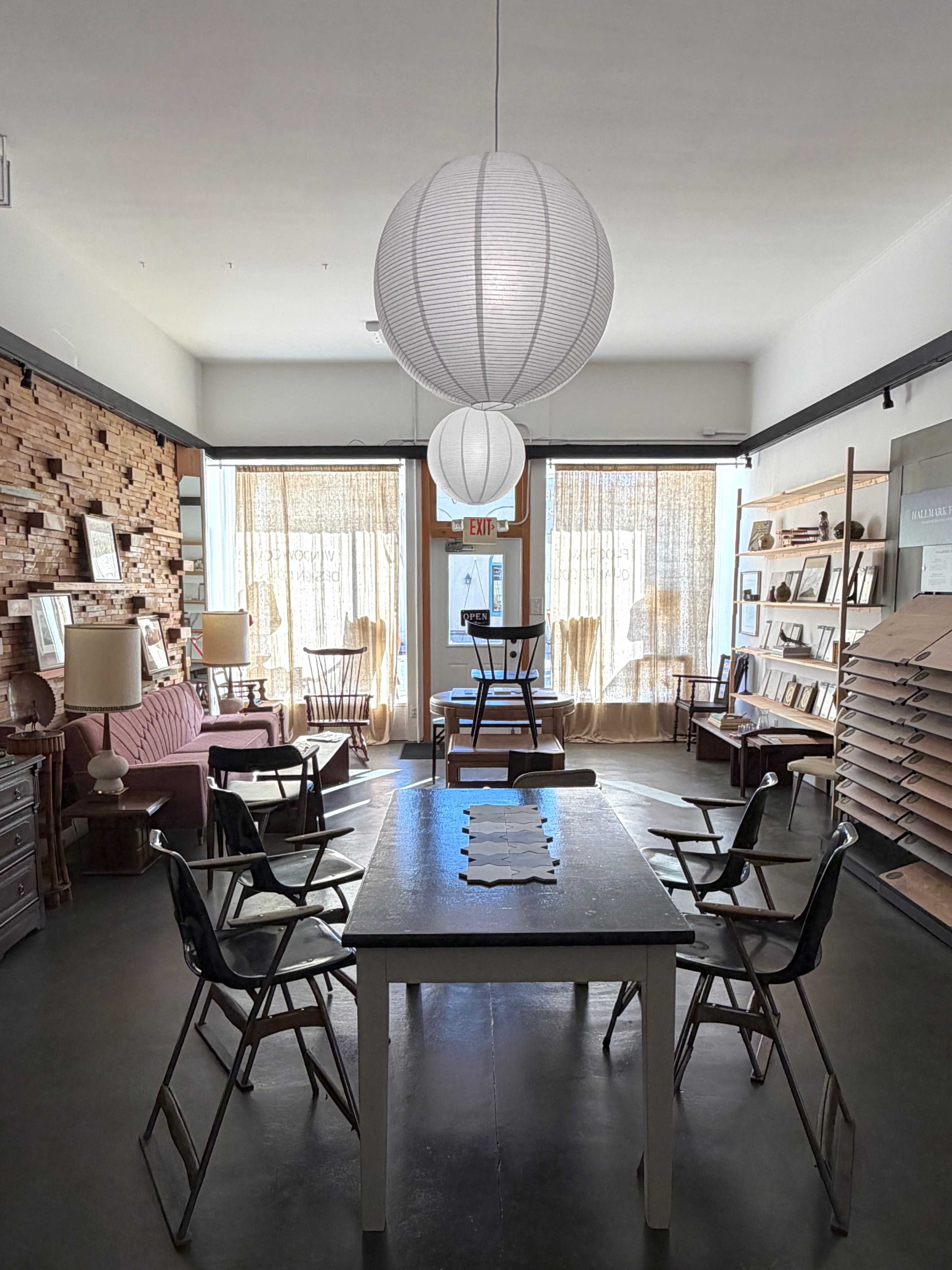 The image shows a spacious interior of a room with a central table surrounded by chairs, illuminated by three hanging lamps, and featuring a wall of wood paneling and shelves with various items.