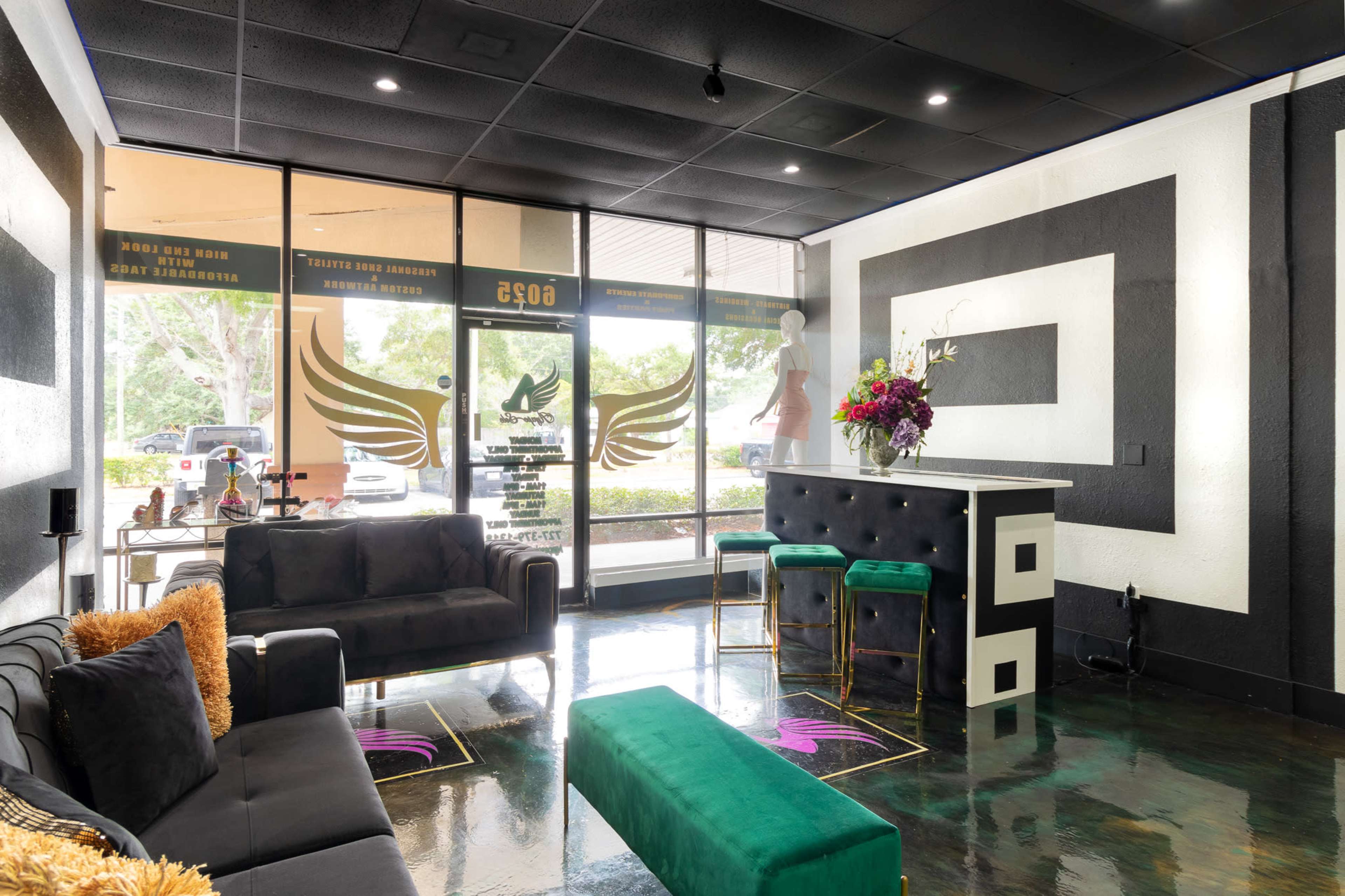The interior of a modern salon features black sofas, a green bench, a stylish reception area with high stools, and large windows showcasing outdoor views.
