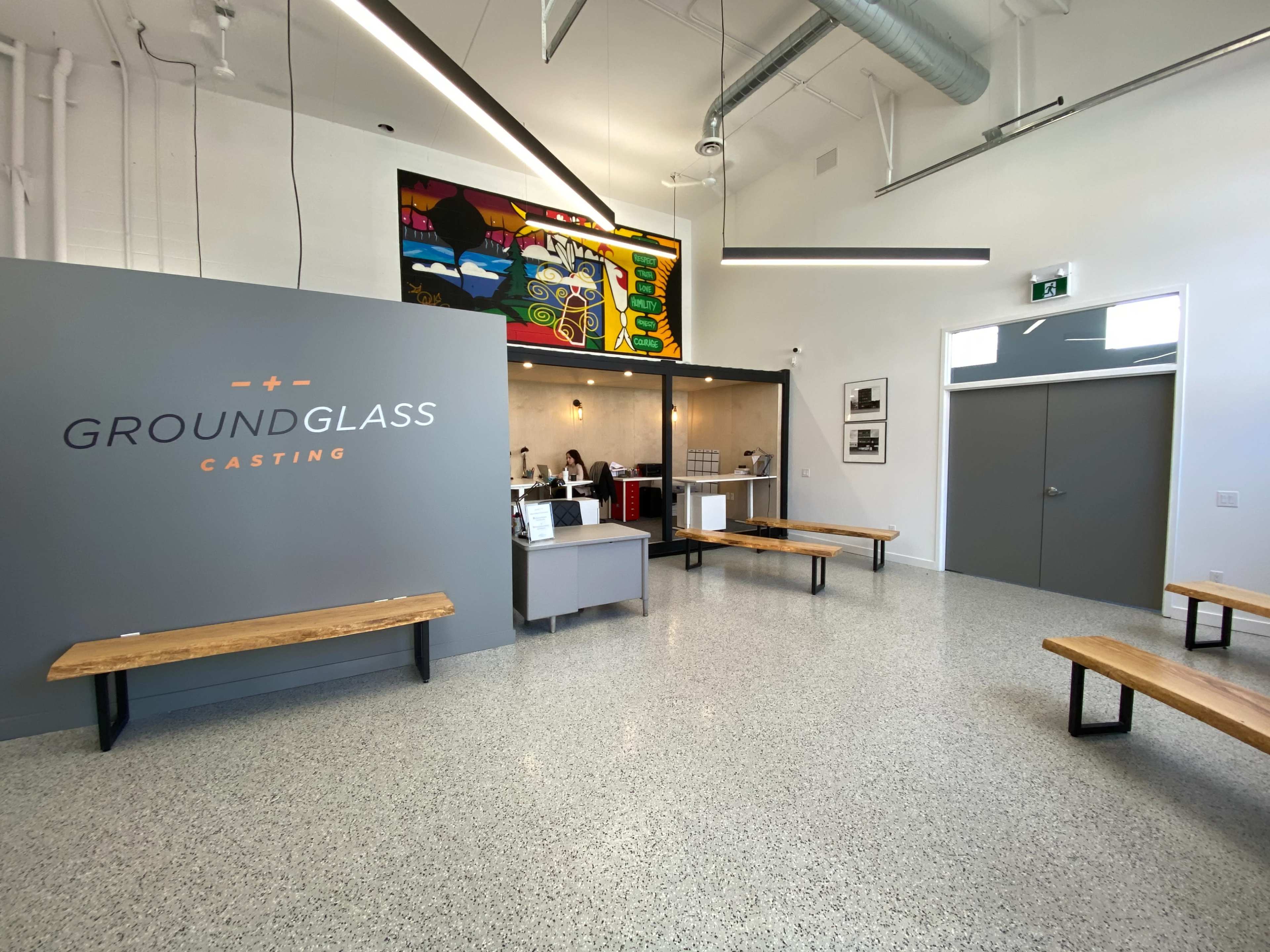 A modern reception area for "Ground Glass Casting," featuring a gray wall with the company name, wooden benches, and a colorful mural in the background.