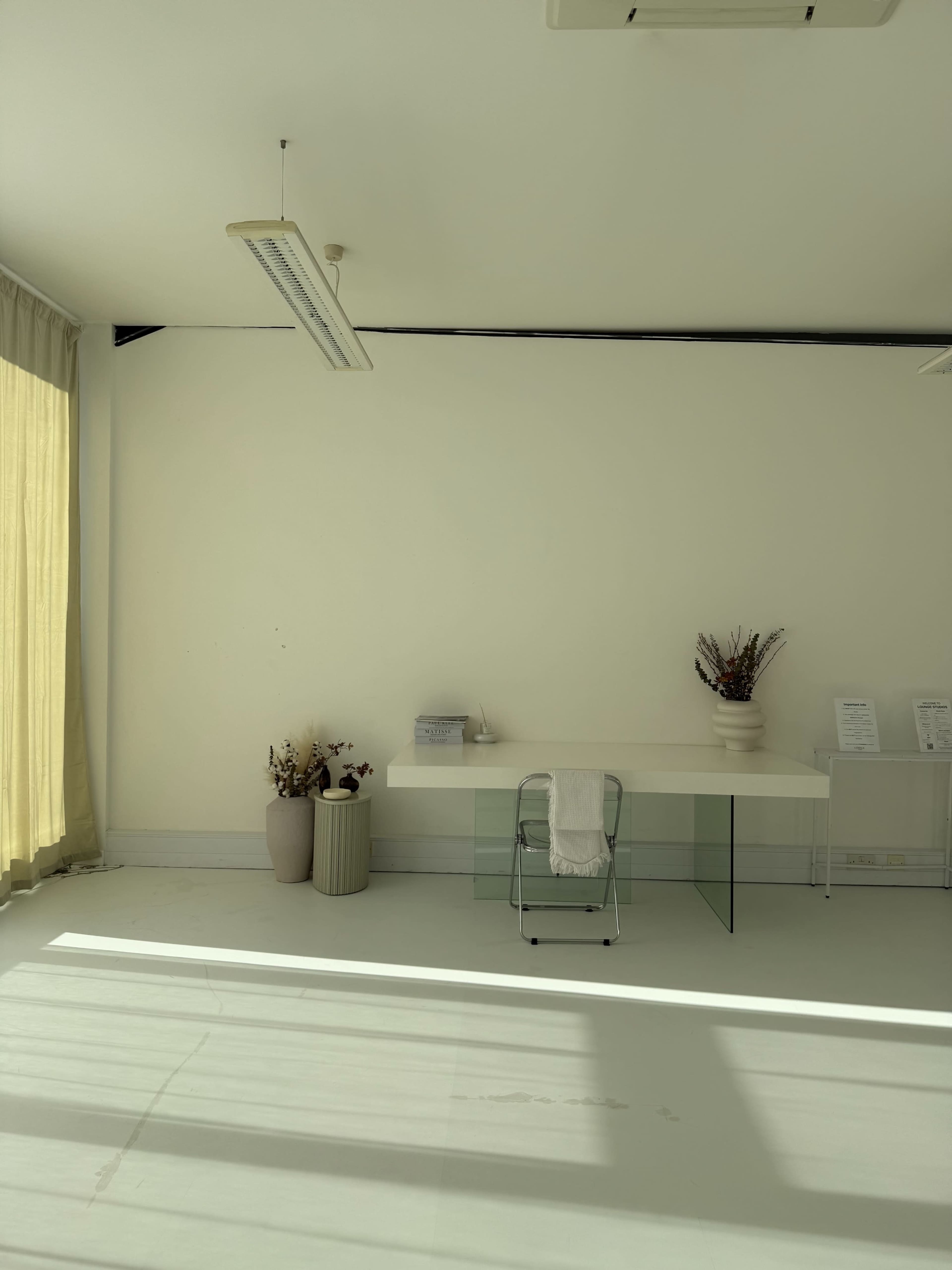 The image shows a minimalistic room with a transparent desk, two potted plants, and large windows casting shadows on the white floor.