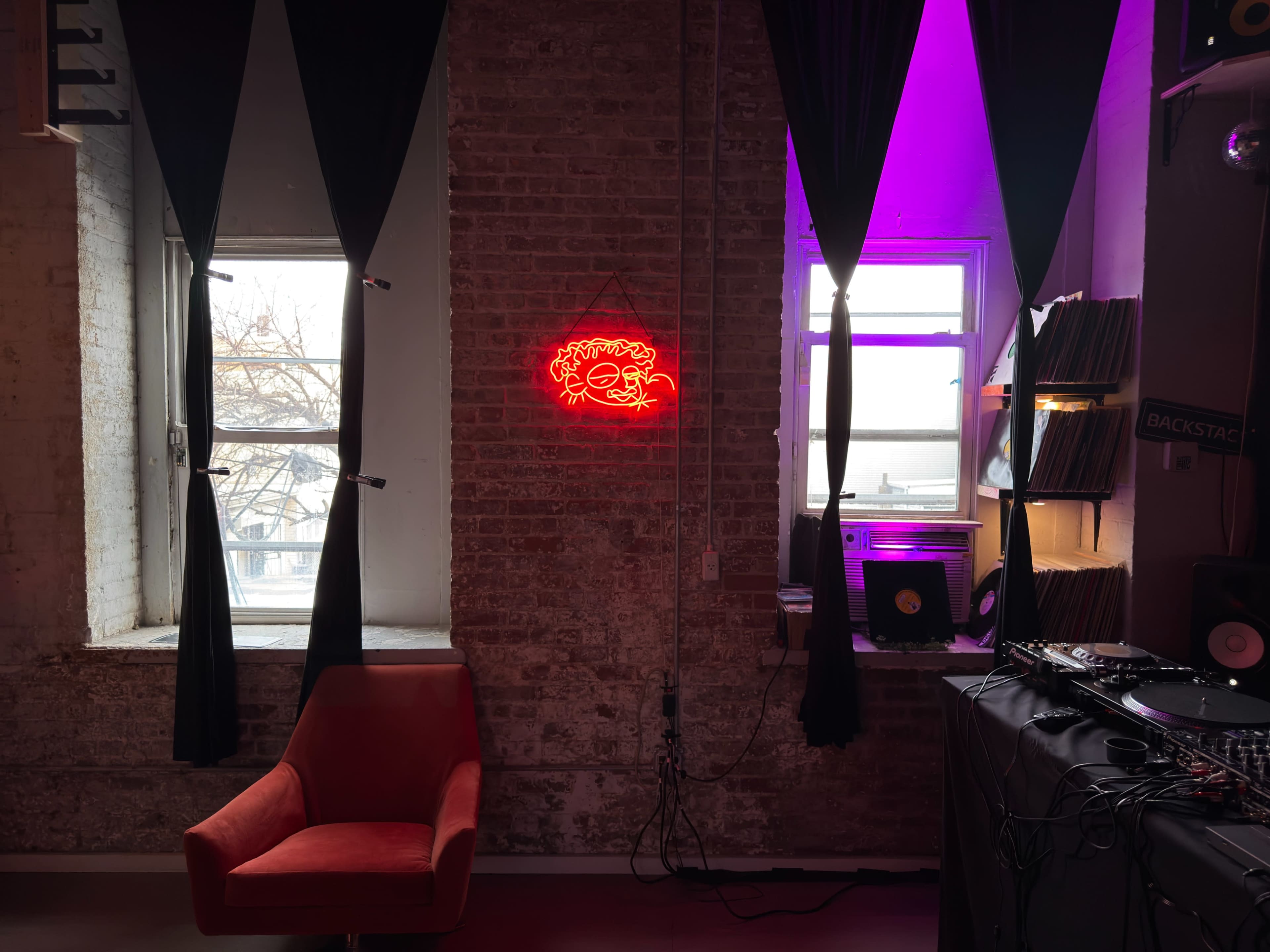 The image shows a room with a red neon sign on a brick wall, two windows draped in black curtains, a red chair, and a setup for DJ equipment.
