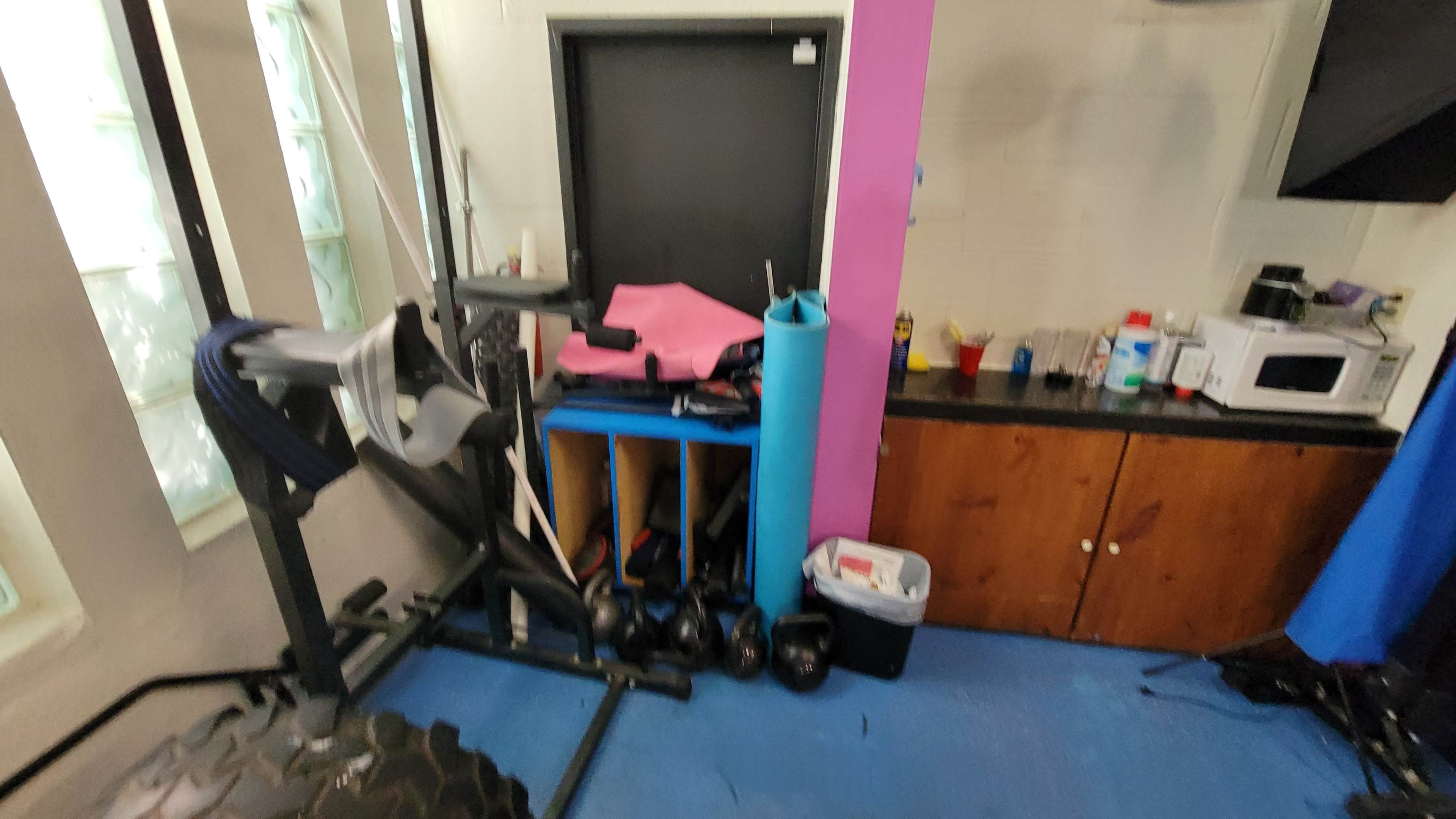 The image shows a corner of a gym with exercise equipment, a stack of mats, a trash can, and a microwave on a countertop.