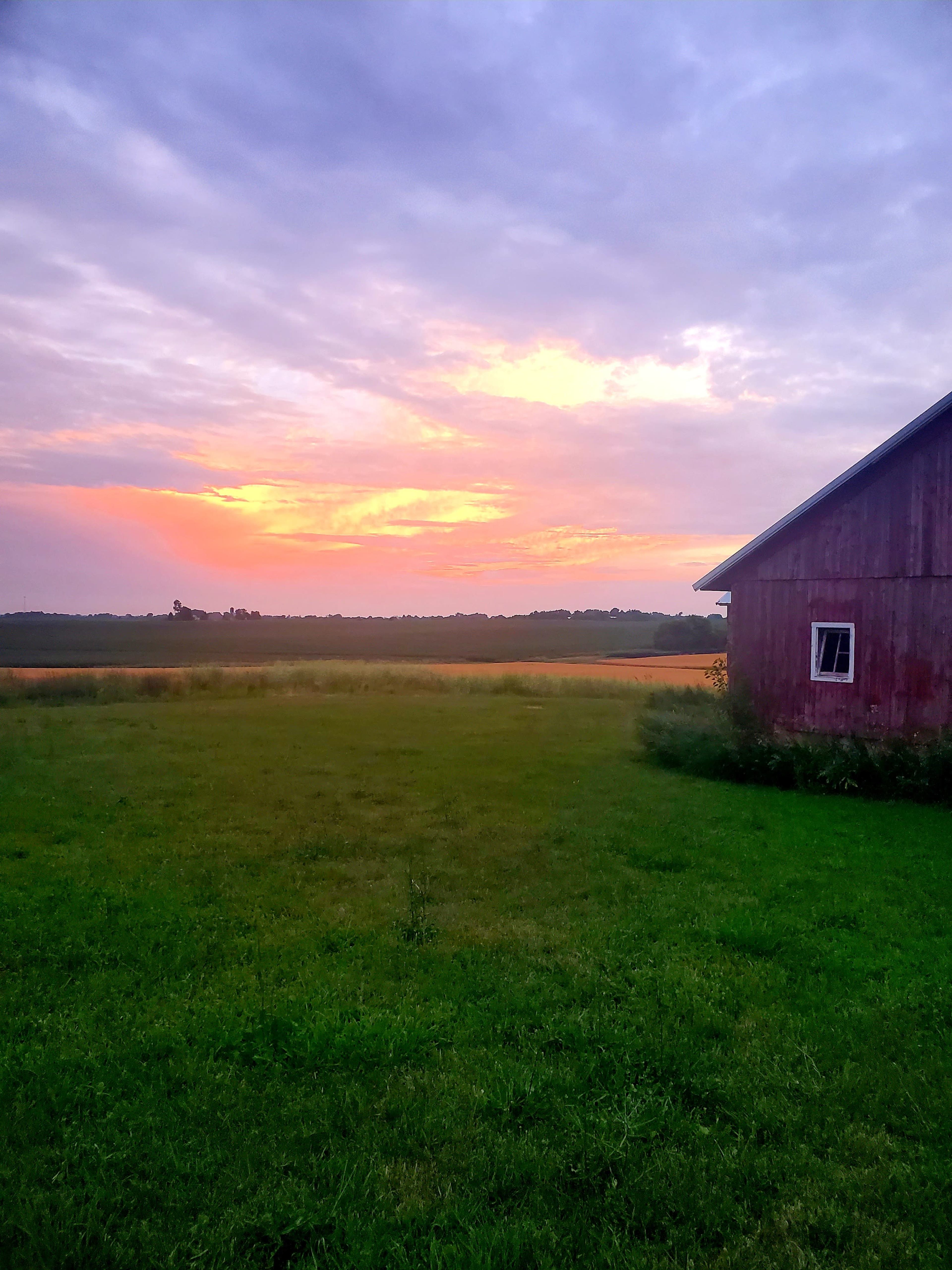 A red barn stands on green grass under a colorful sky at sunset, with fields stretching into the distance.