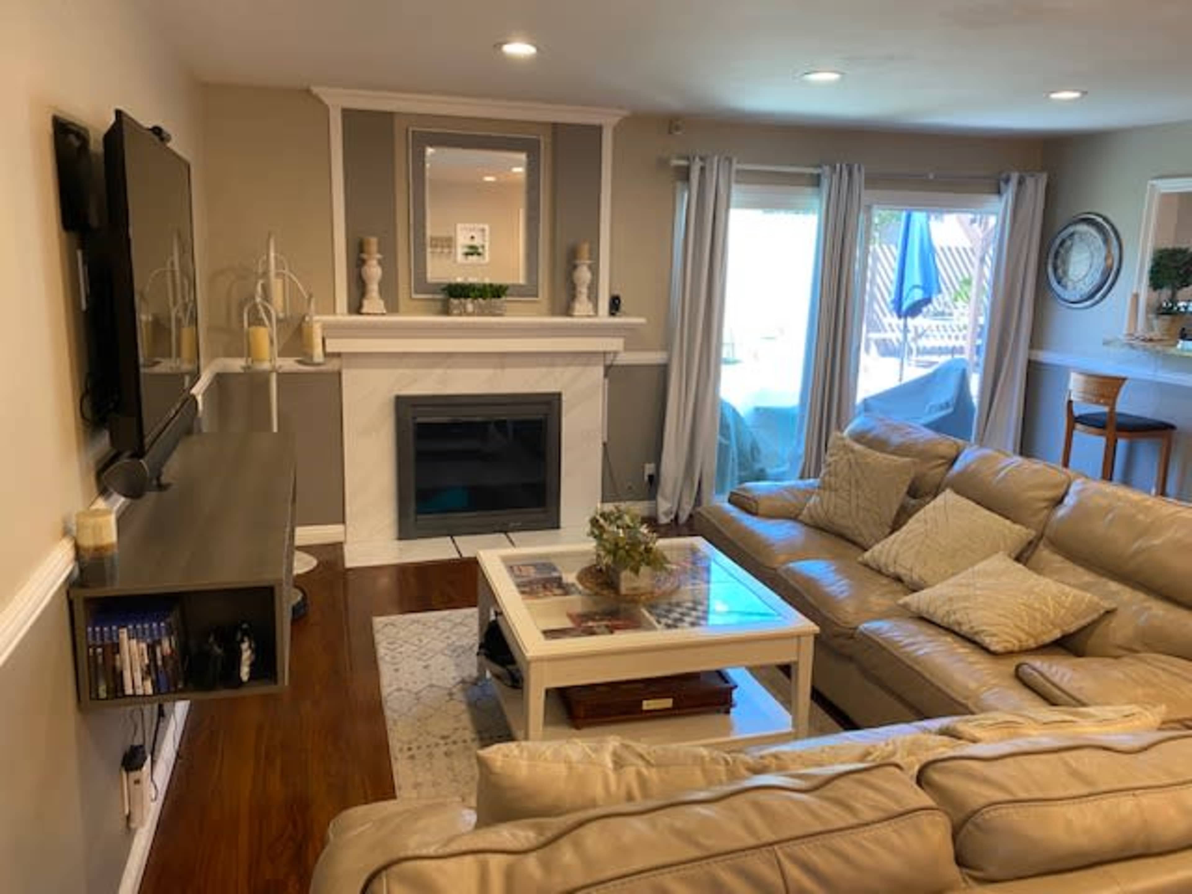 A living room with a cream-colored leather sectional sofa, a coffee table, a wall-mounted TV, and a white marble fireplace, alongside large windows leading to an outdoor patio.