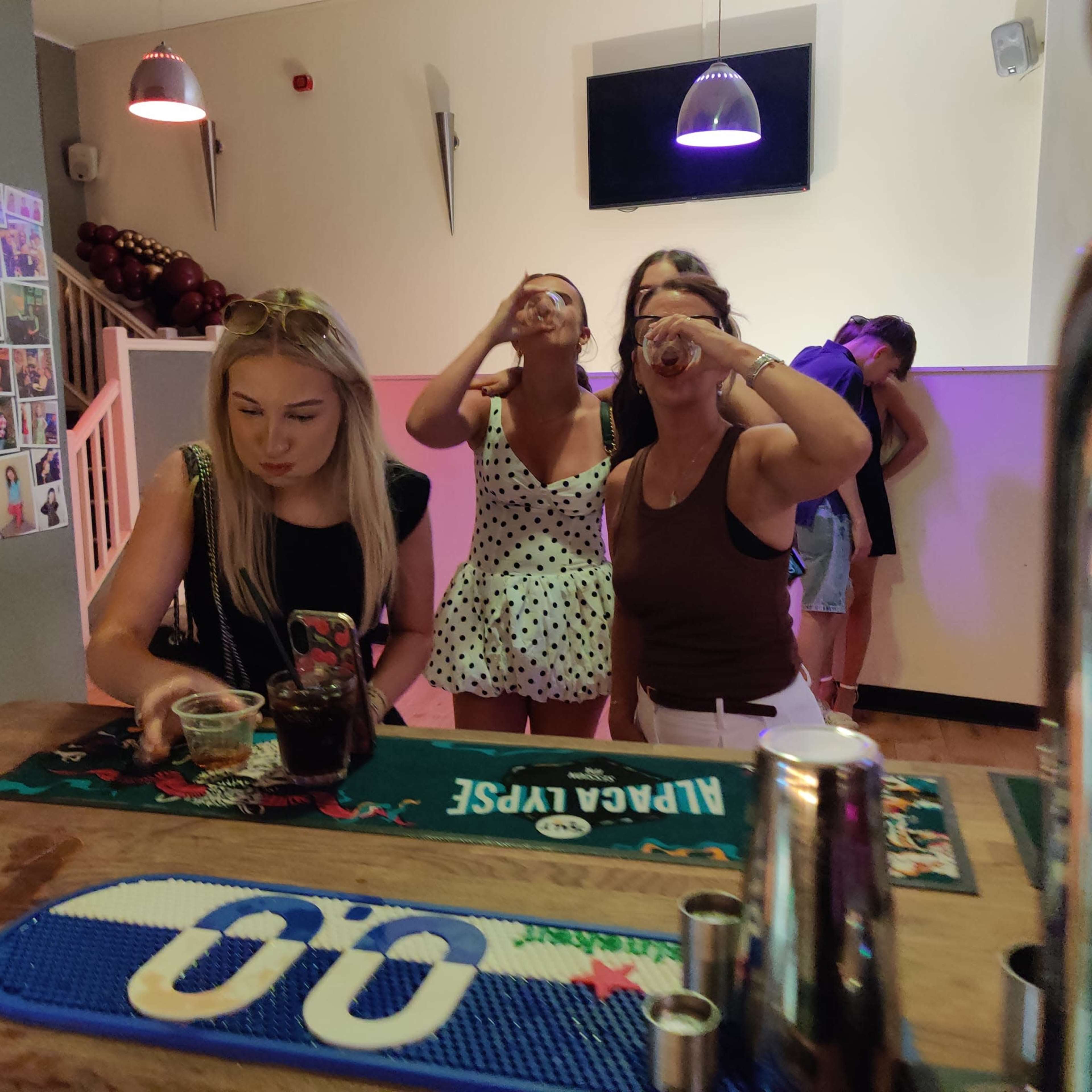A group of three women in a bar are raising their glasses for a toast while another person is visible in the background.