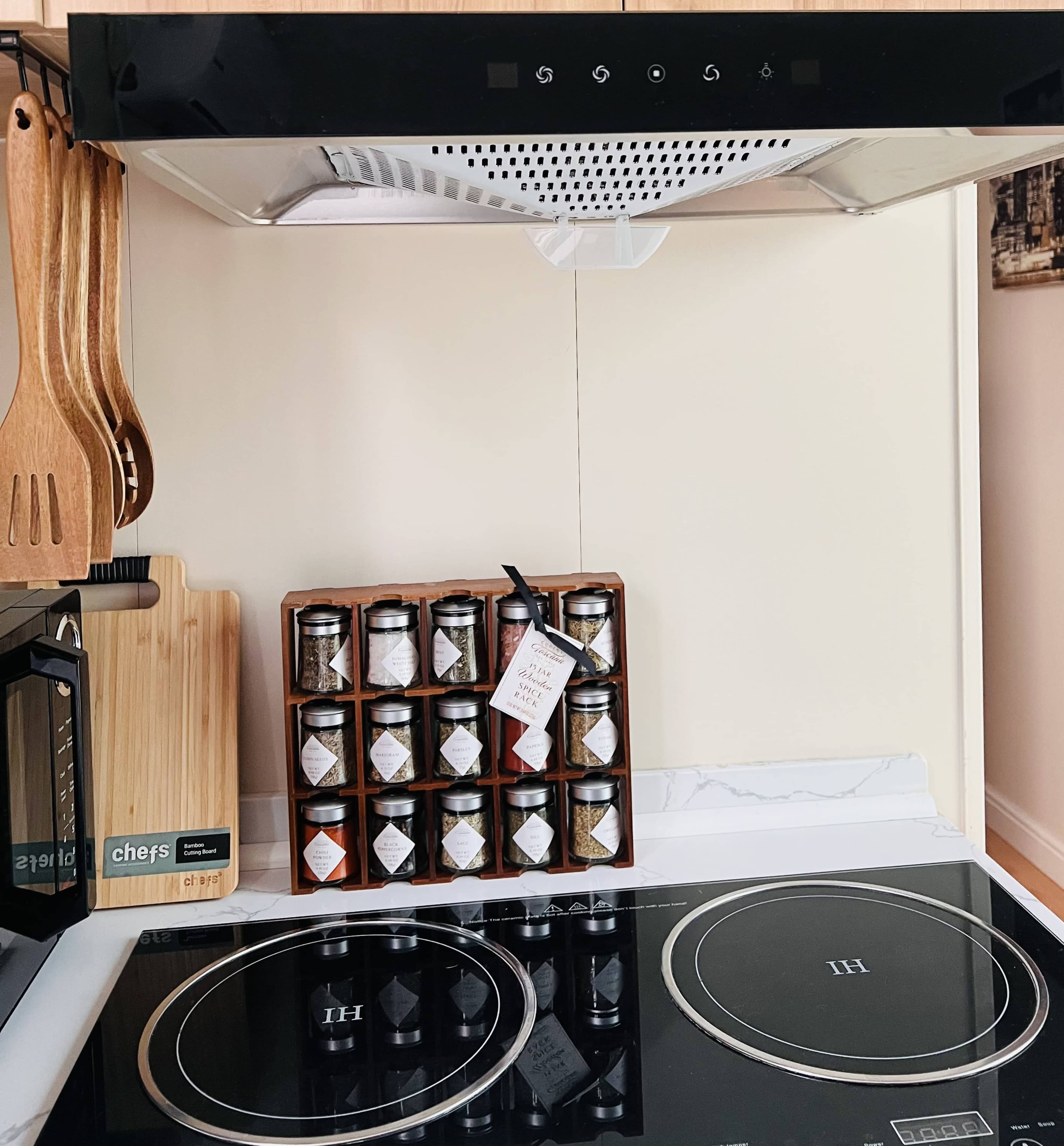 A kitchen countertop with an induction cooktop, a wooden cutting board, wooden utensils, and a rack of spice jars labeled with tags.