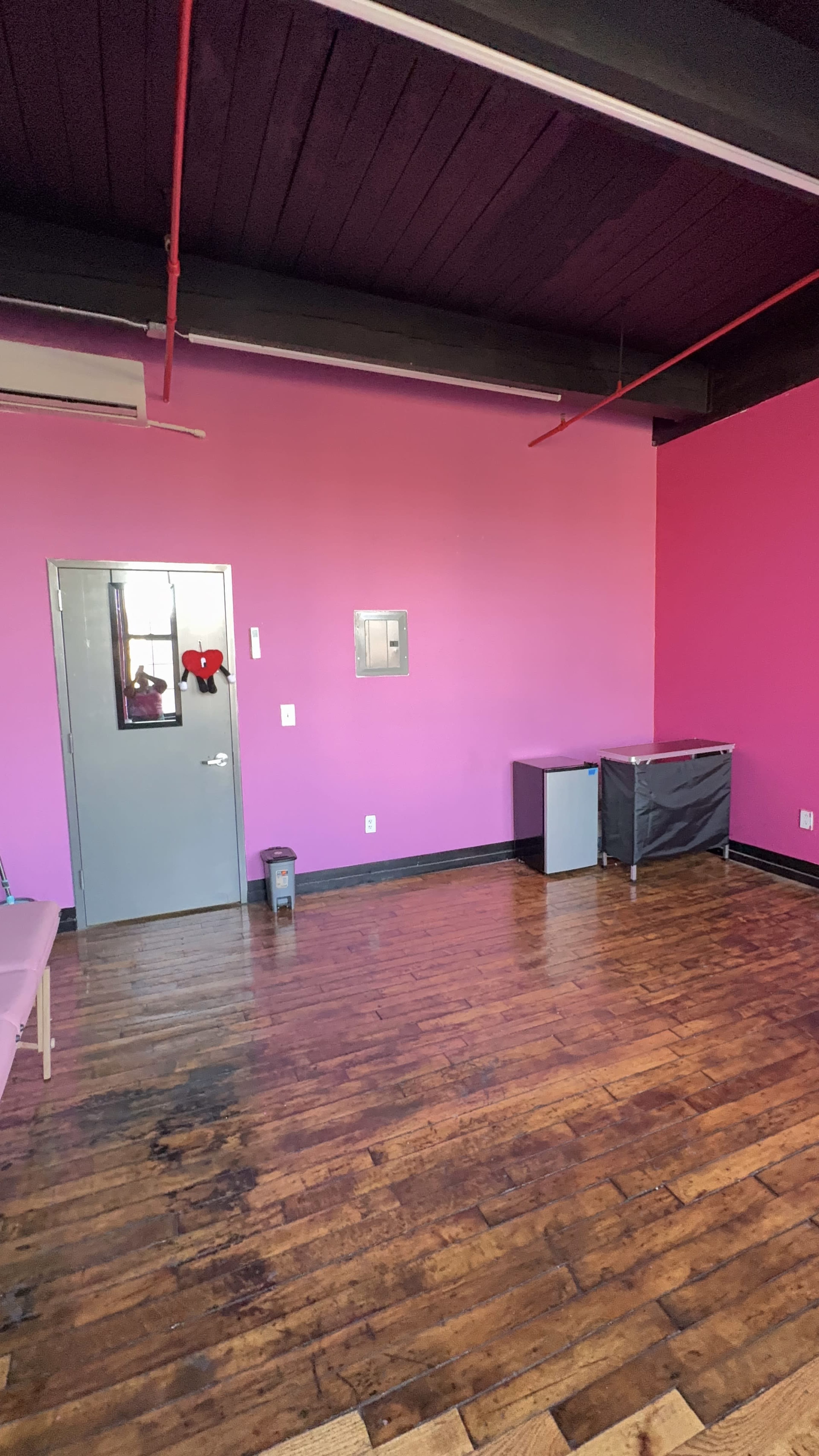 The image shows a room with pink walls, a wooden floor, a gray door, and minimal furniture including a trash can and storage unit.