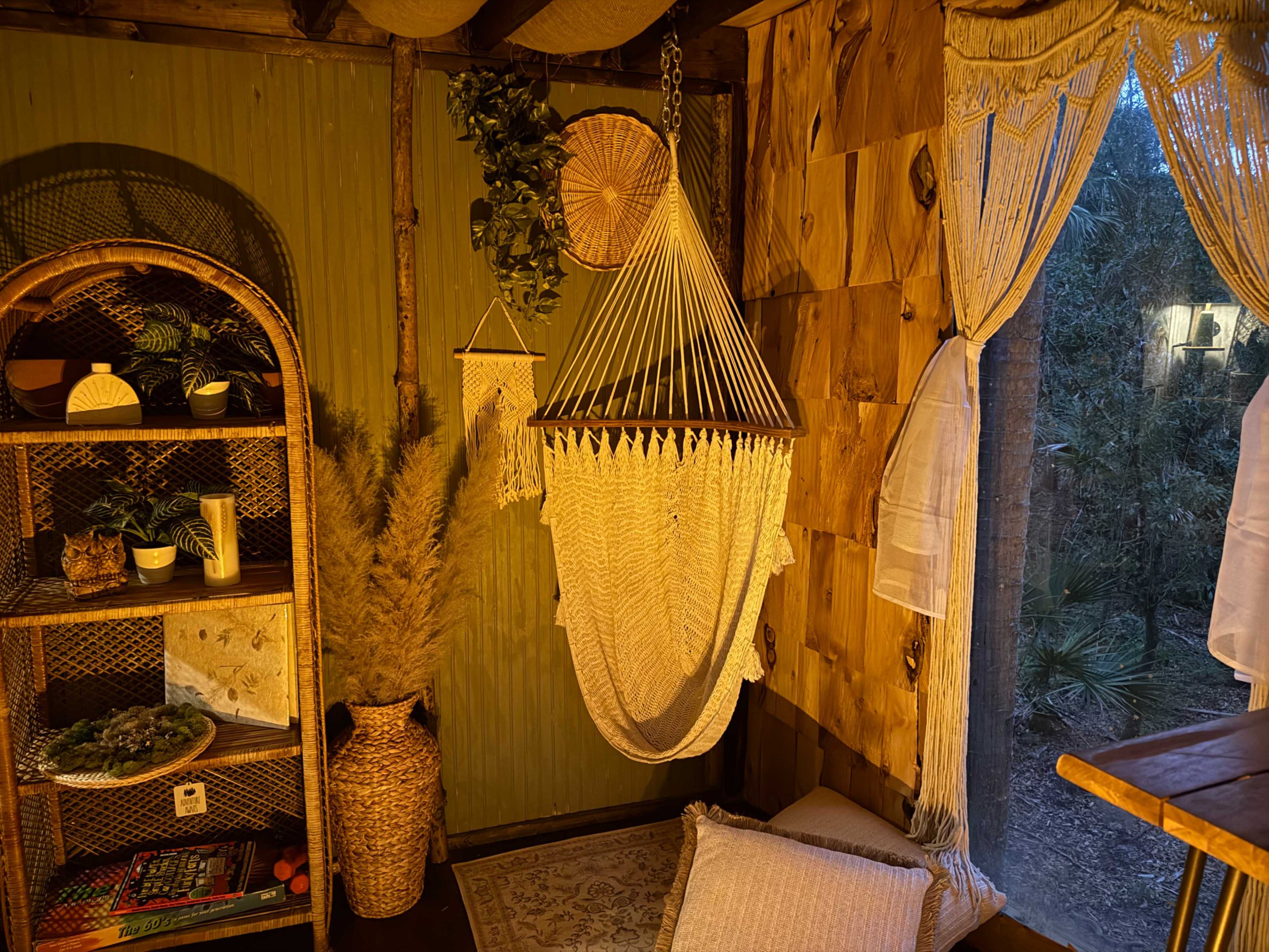 The image shows a cozy, softly lit interior featuring a hanging macramé chair, a woven bookshelf filled with decor, and natural elements like potted plants and dried grasses.