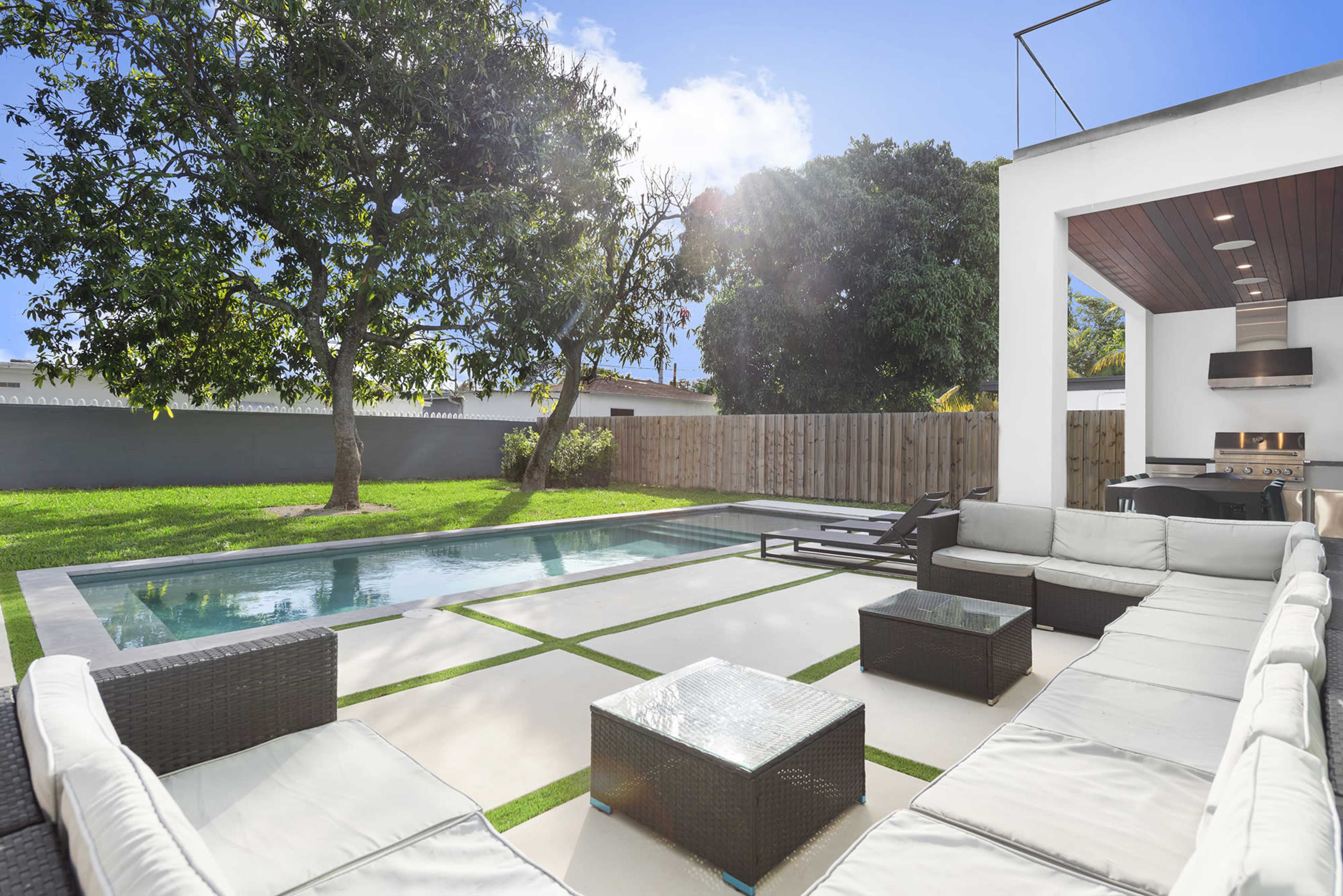 A modern backyard features a swimming pool, outdoor seating area with sectional sofas, and a barbecue station surrounded by a well-maintained lawn and landscaping.