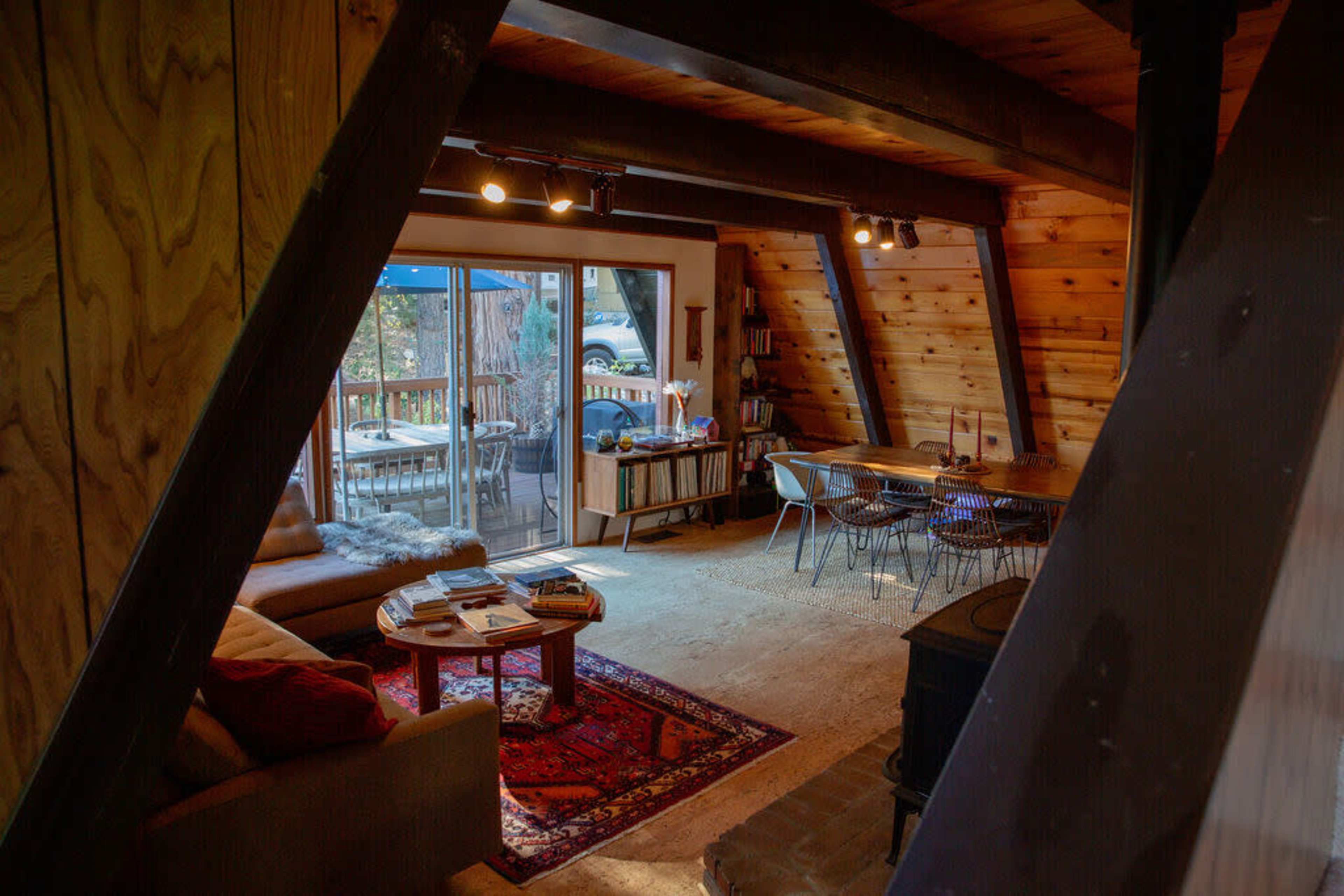 A spacious, rustic living area with wooden beams, a large window overlooking a deck, a seating area with a couch, and a dining table with chairs.