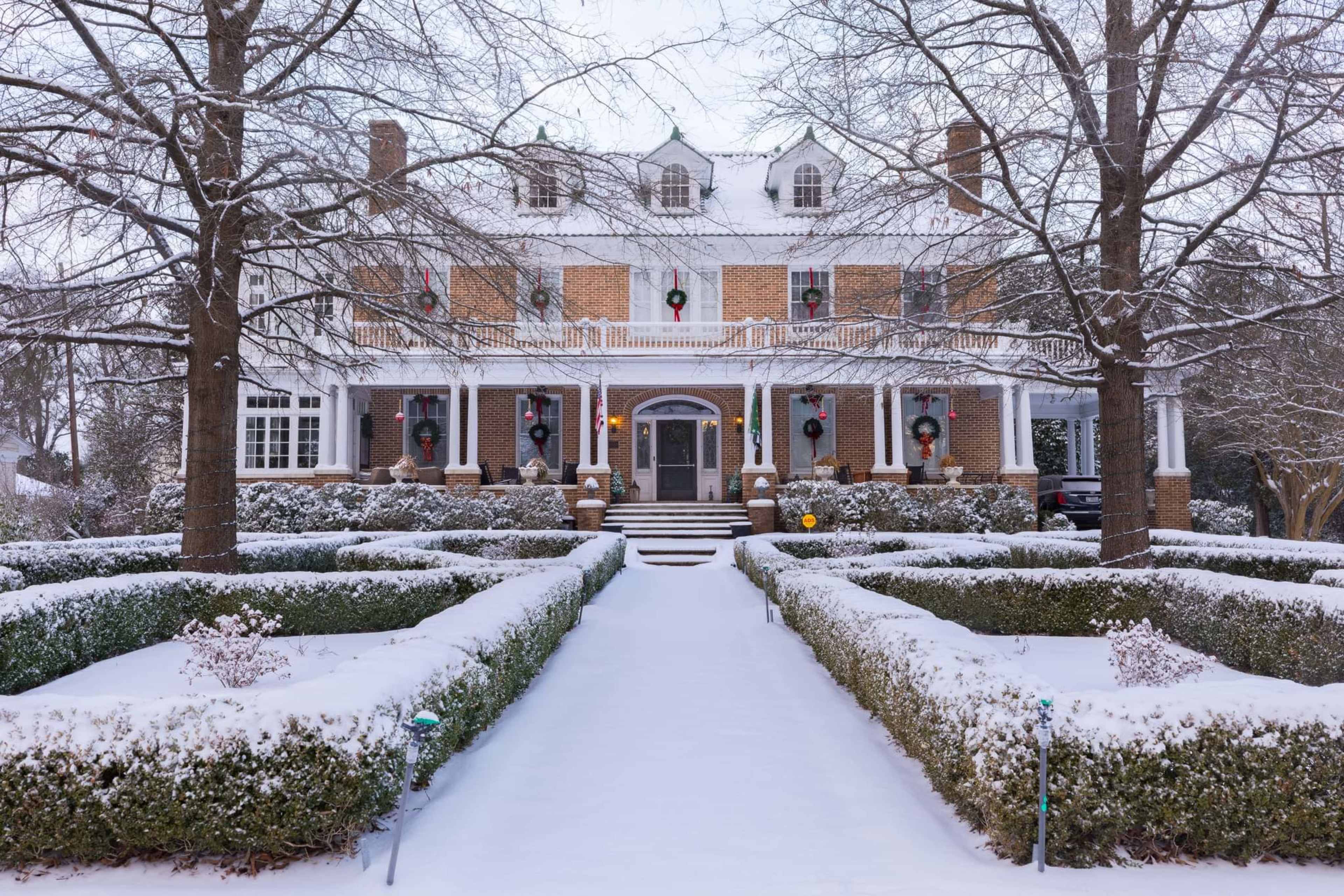 A large brick house with a white porch is surrounded by snow-covered shrubs and a pathway leading to its entrance, with festive wreaths decorating the windows.