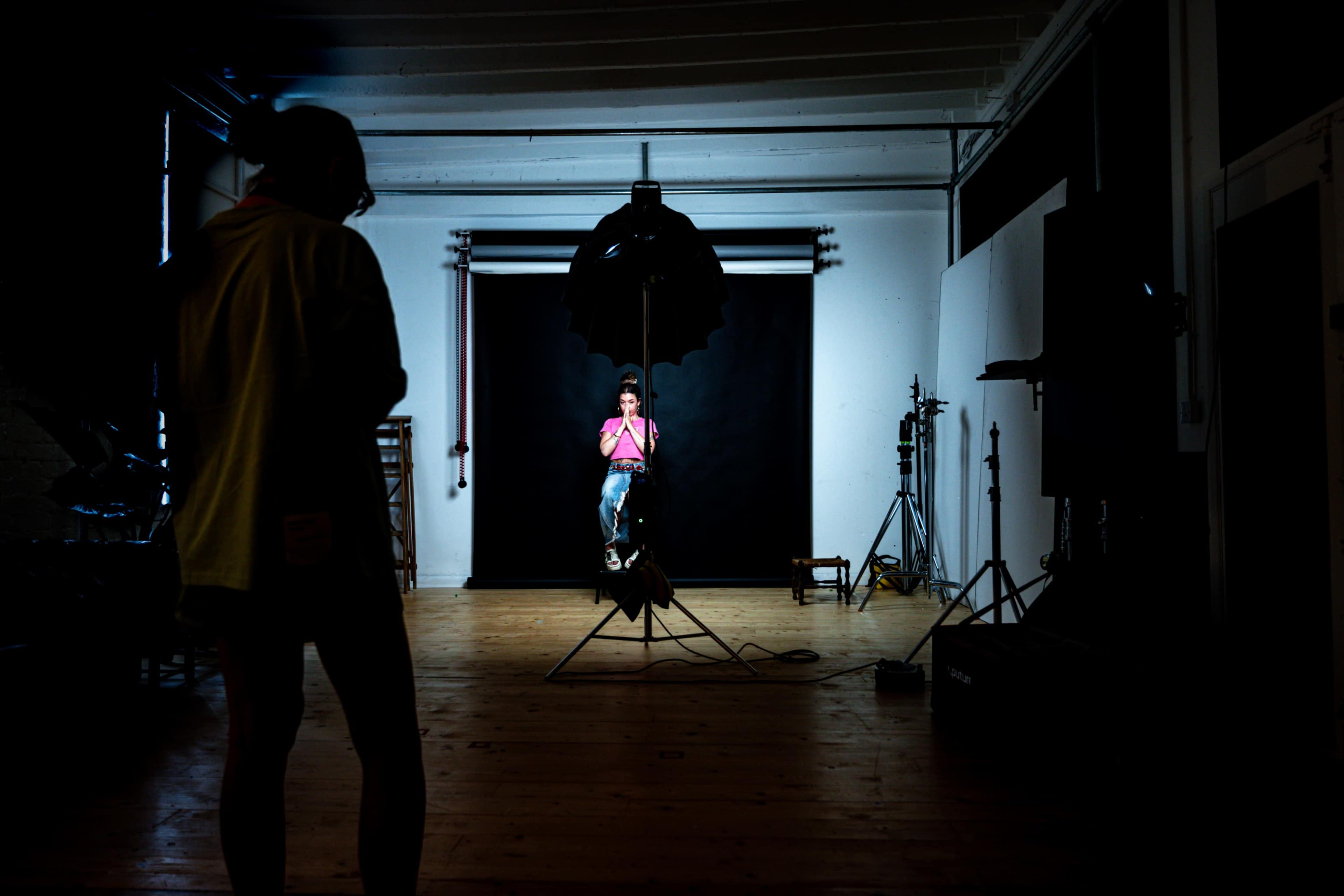 A photographer stands in a dimly lit studio while a model poses in front of a black backdrop.