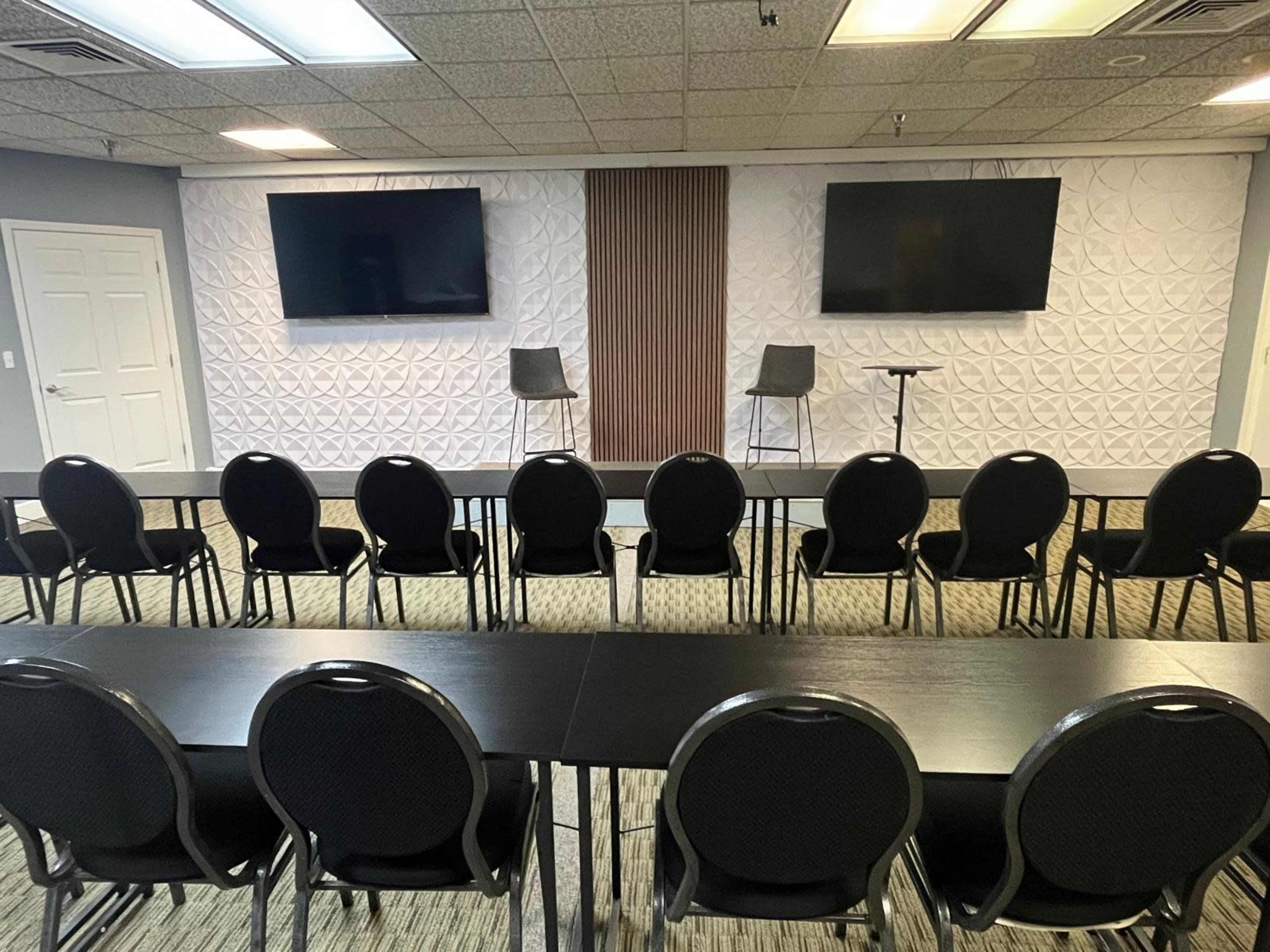 The image shows a conference room set up with two empty chairs on a platform, flanked by two large screens, and rows of black chairs facing a long table.