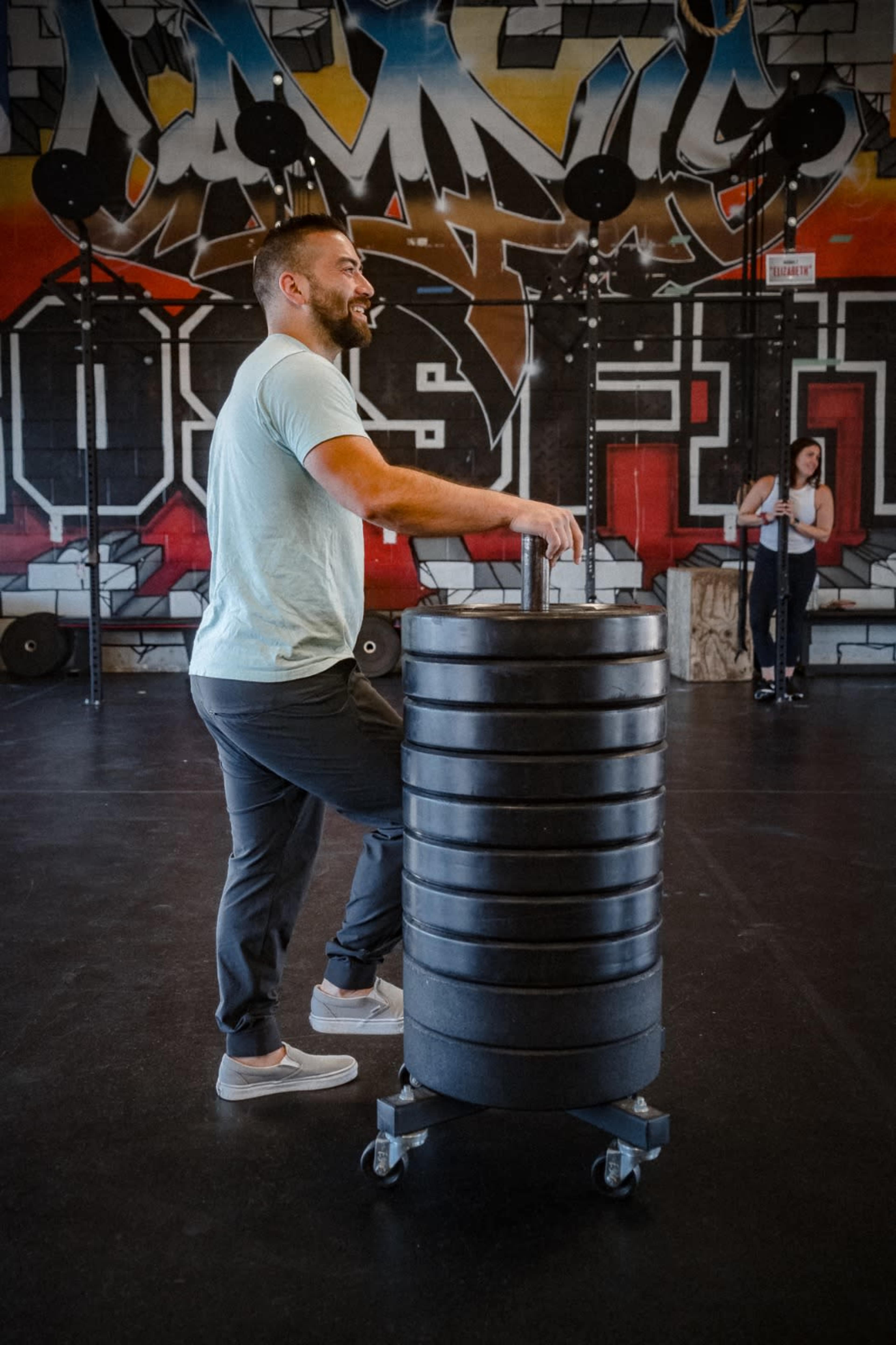 A man in a light shirt stands beside a wheeled weight stack in a gym decorated with colorful graffiti.