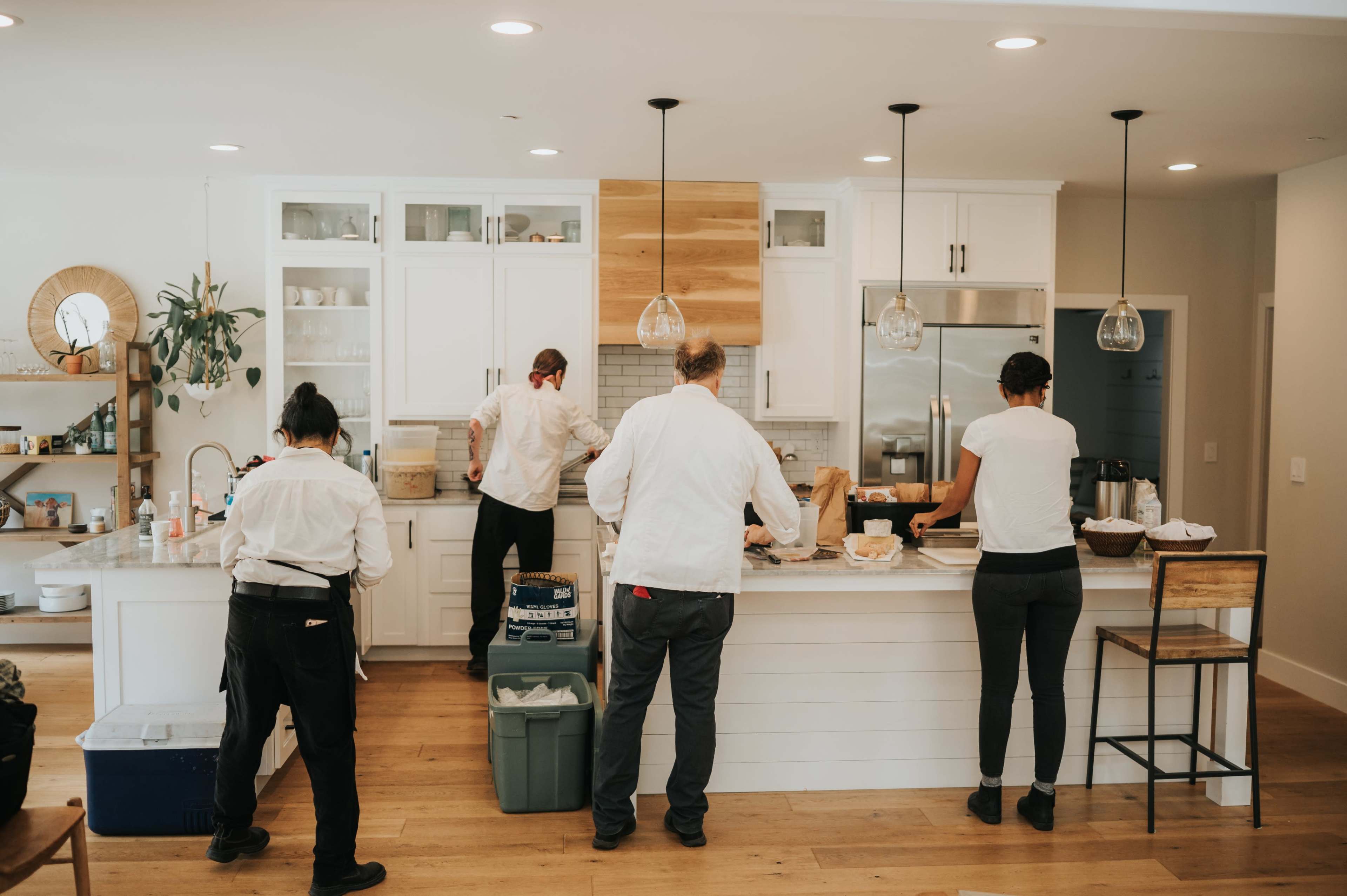 Four individuals in white shirts prepare food in a modern kitchen with a central island and wooden accents.