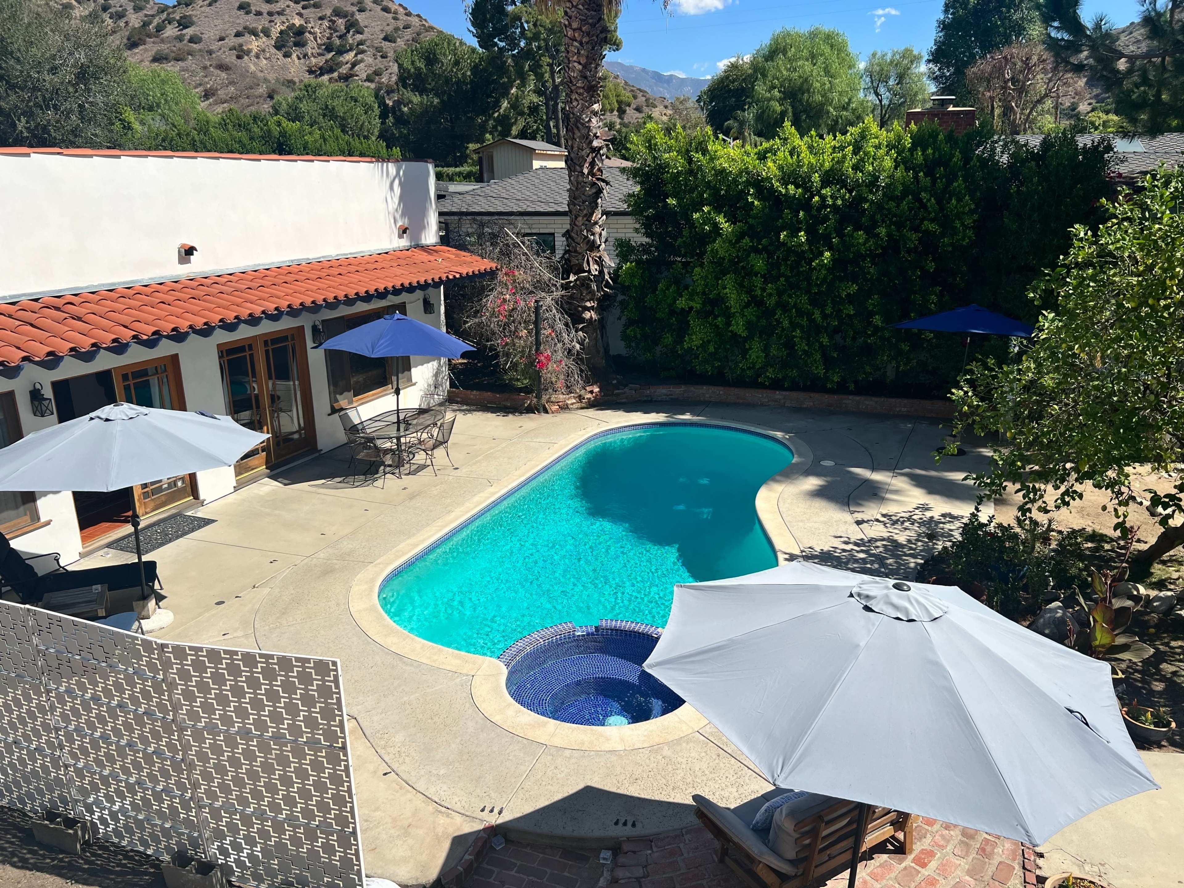 The image shows a backyard with a swimming pool, surrounded by lounge chairs and umbrellas, amid a landscape of trees and mountains.