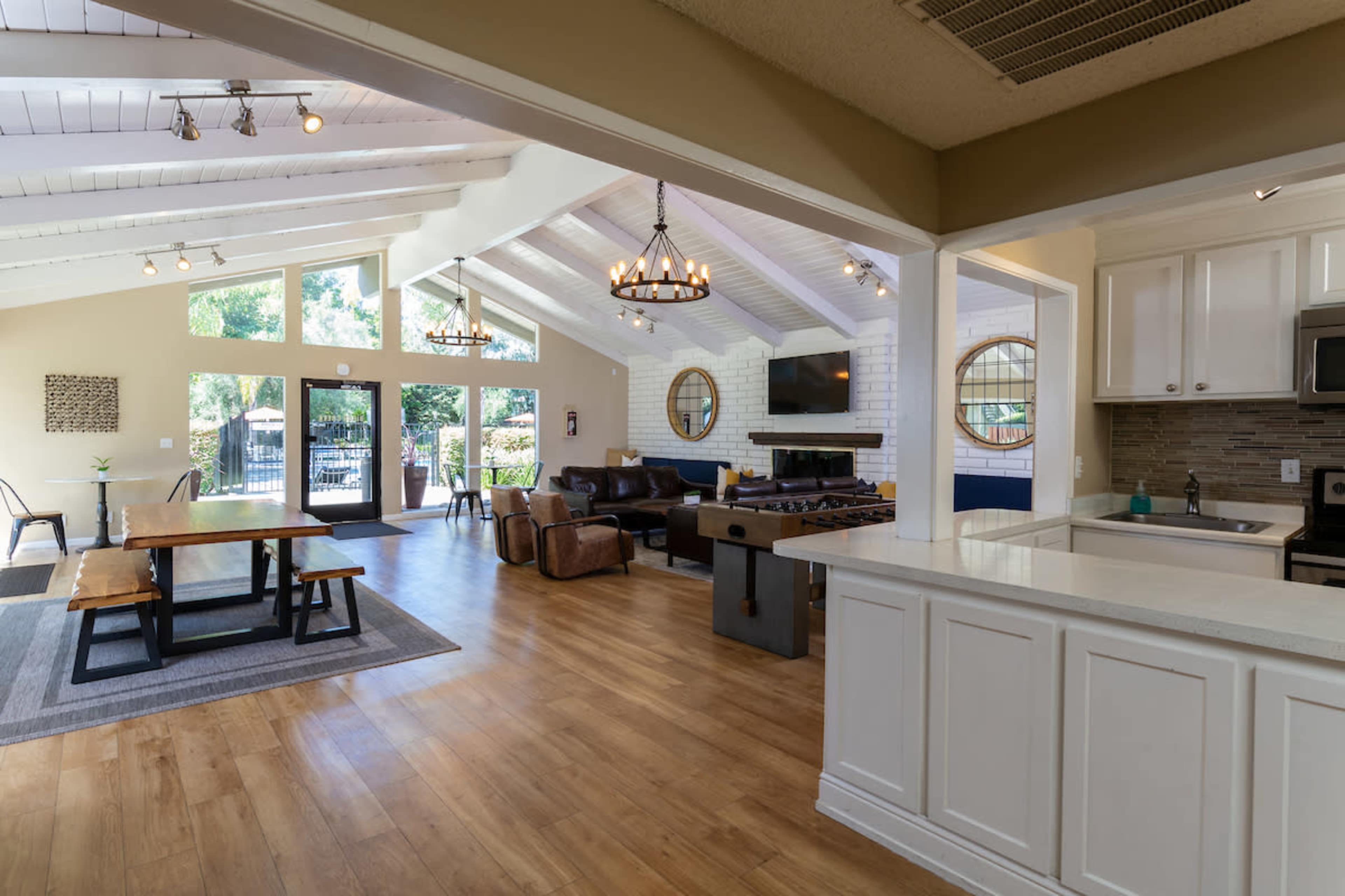 The image shows an open-concept living space with a kitchen, dining area featuring a large table, and a cozy seating arrangement adjacent to windows that overlook an outdoor patio.