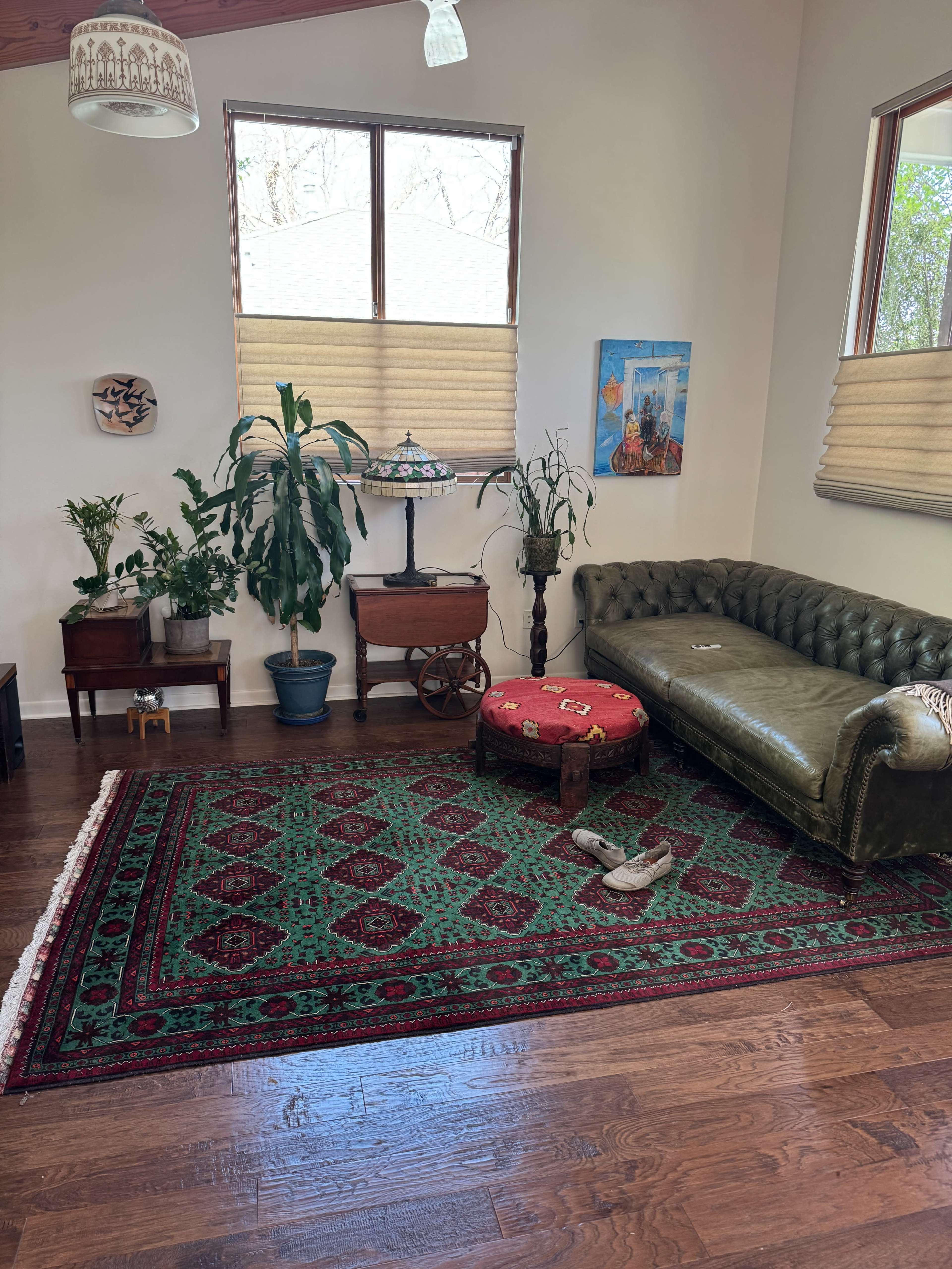 The image shows a spacious living room with a green leather couch, a round table, potted plants, and a large patterned rug on a wooden floor.