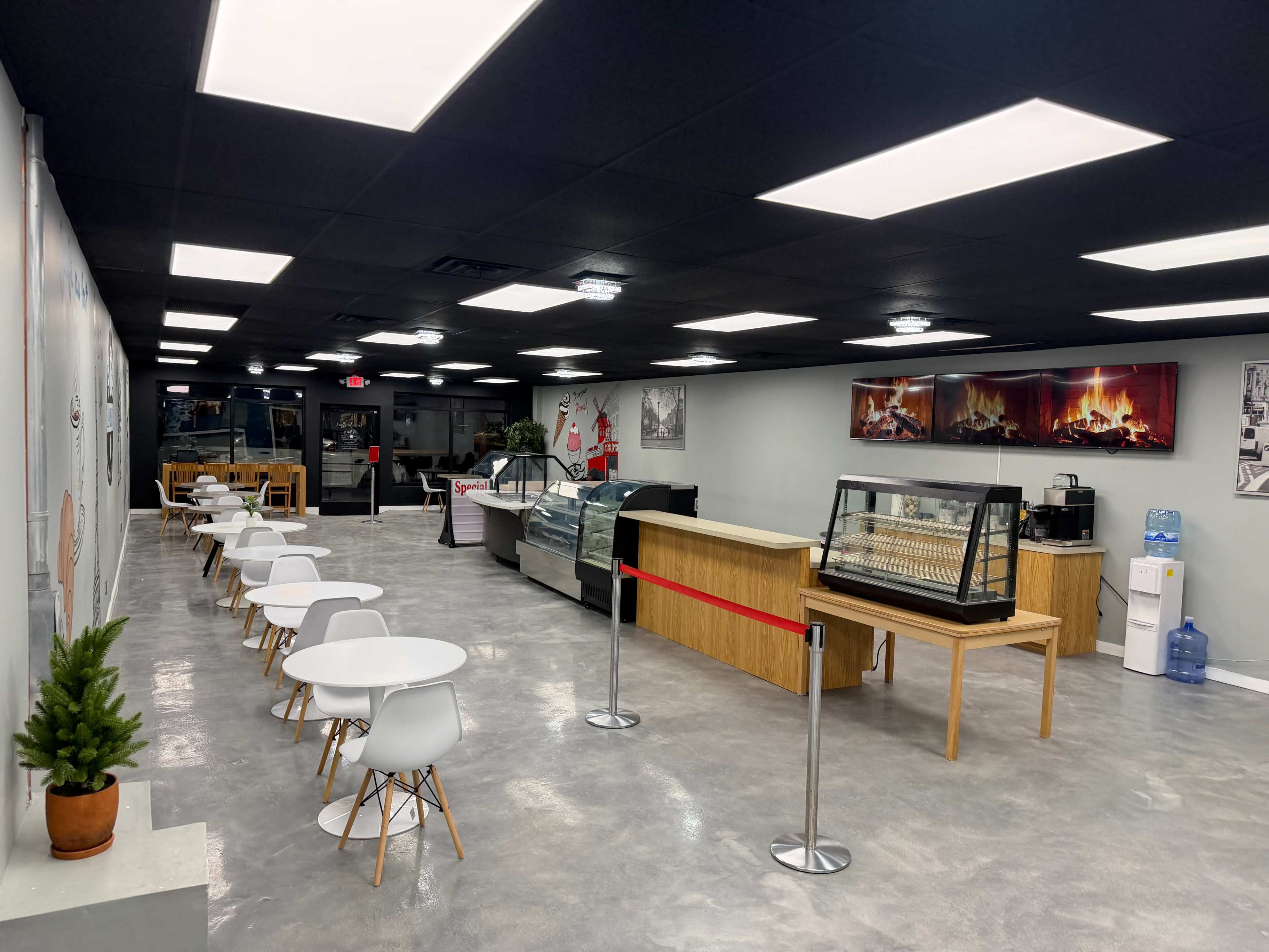 The image shows a modern café interior with white tables and chairs, a display case, and a counter area, illuminated by bright ceiling lights.